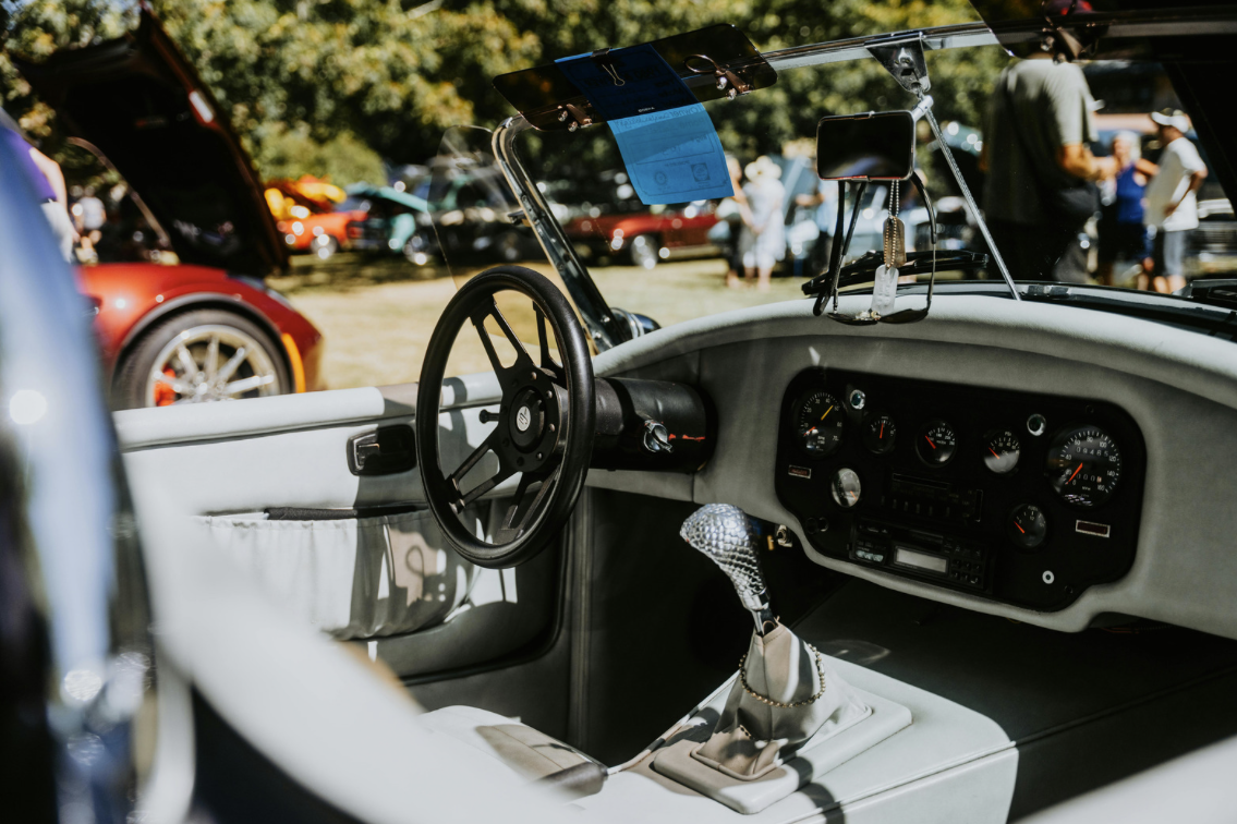 Interior of a classic car with a black steering wheel, gear shift with a sparkle cover, and analog gauges on the dashboard, at a car show with several people and cars in the background.