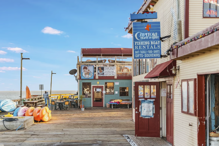 View of a fishing boat rentals shop on a wooden pier, with colorful buoys, outdoor seating, and a clear blue sky.