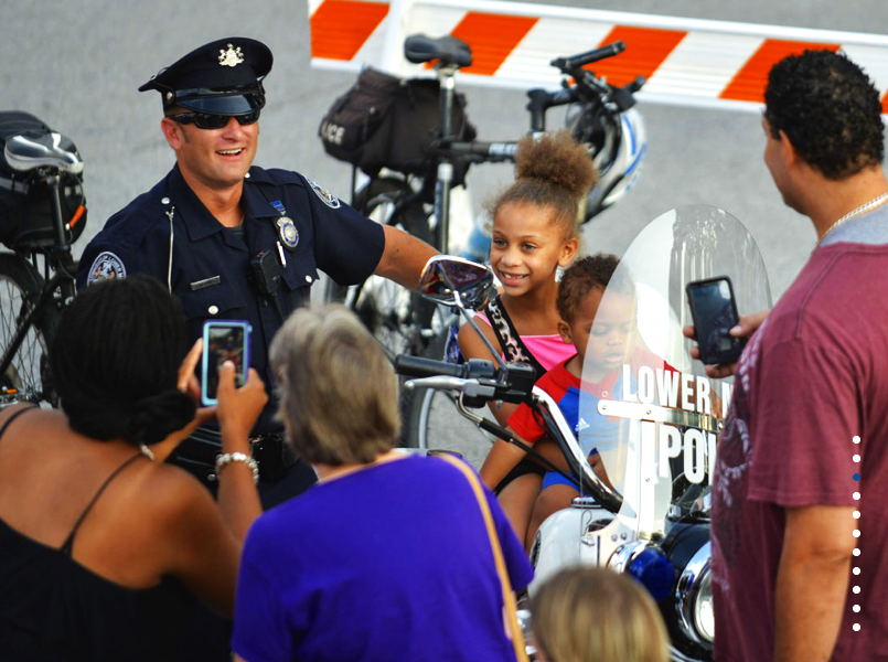 A police officer in uniform is smiling and talking to a group of people gathered around a police motorcycle, with children and adults taking pictures.
