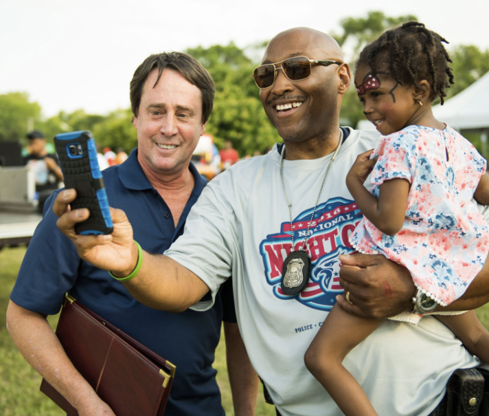 Two men and a young girl taking a selfie outdoors at a community event, with tents and trees in the background.
