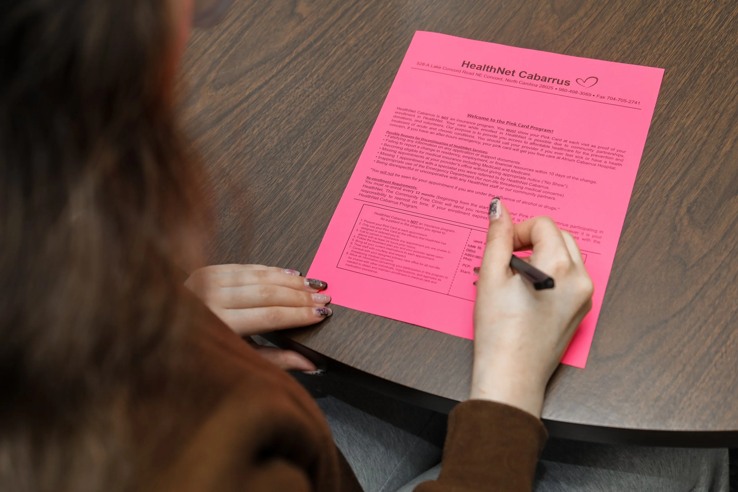 A person with manicured nails is writing on a pink health pamphlet at a wooden table.