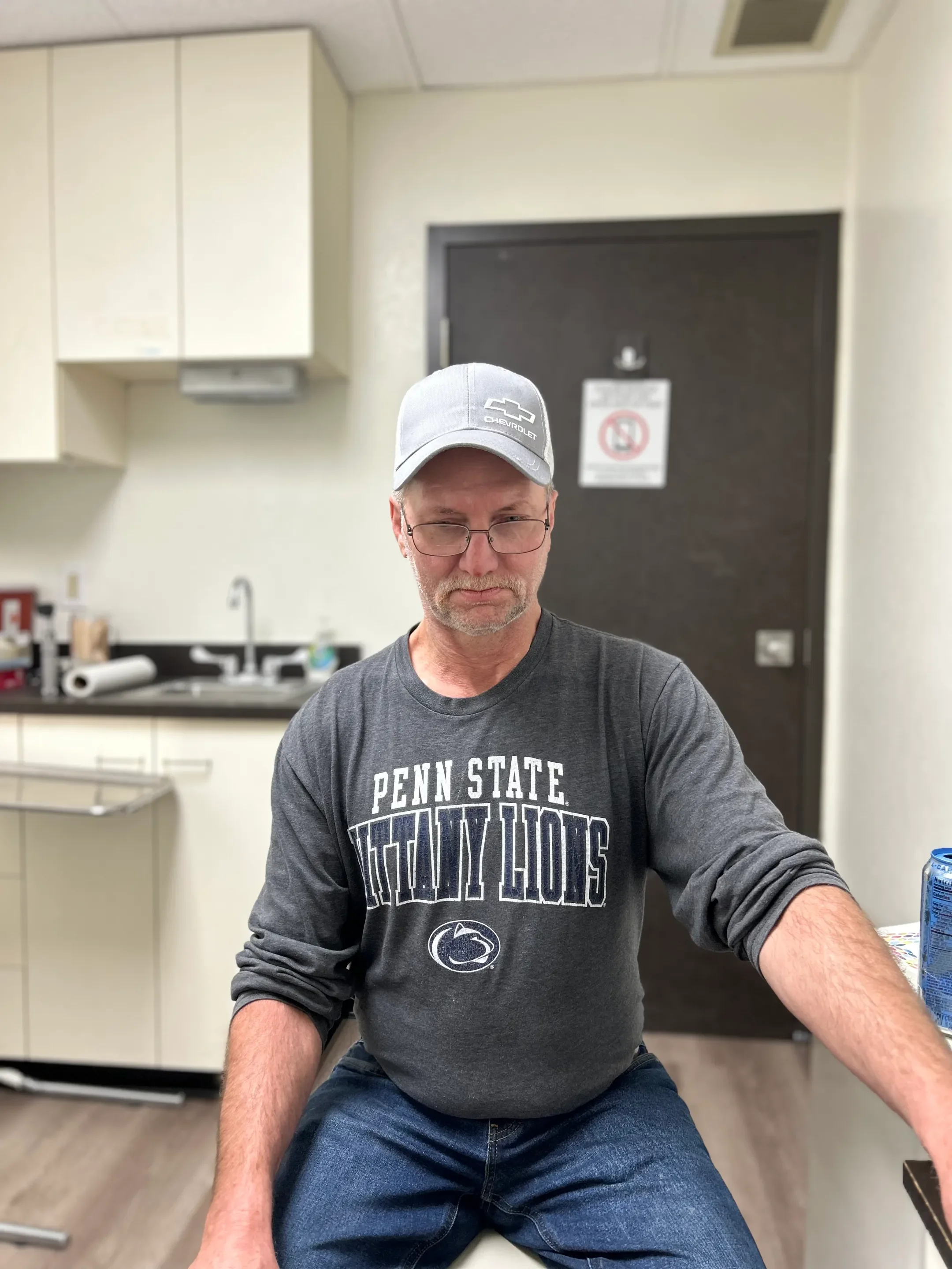 A man wearing glasses, a Penn State Nittany Lions sweatshirt, and a light gray Chevrolet baseball cap sitting in a room with beige cabinets and a dark door in the background.