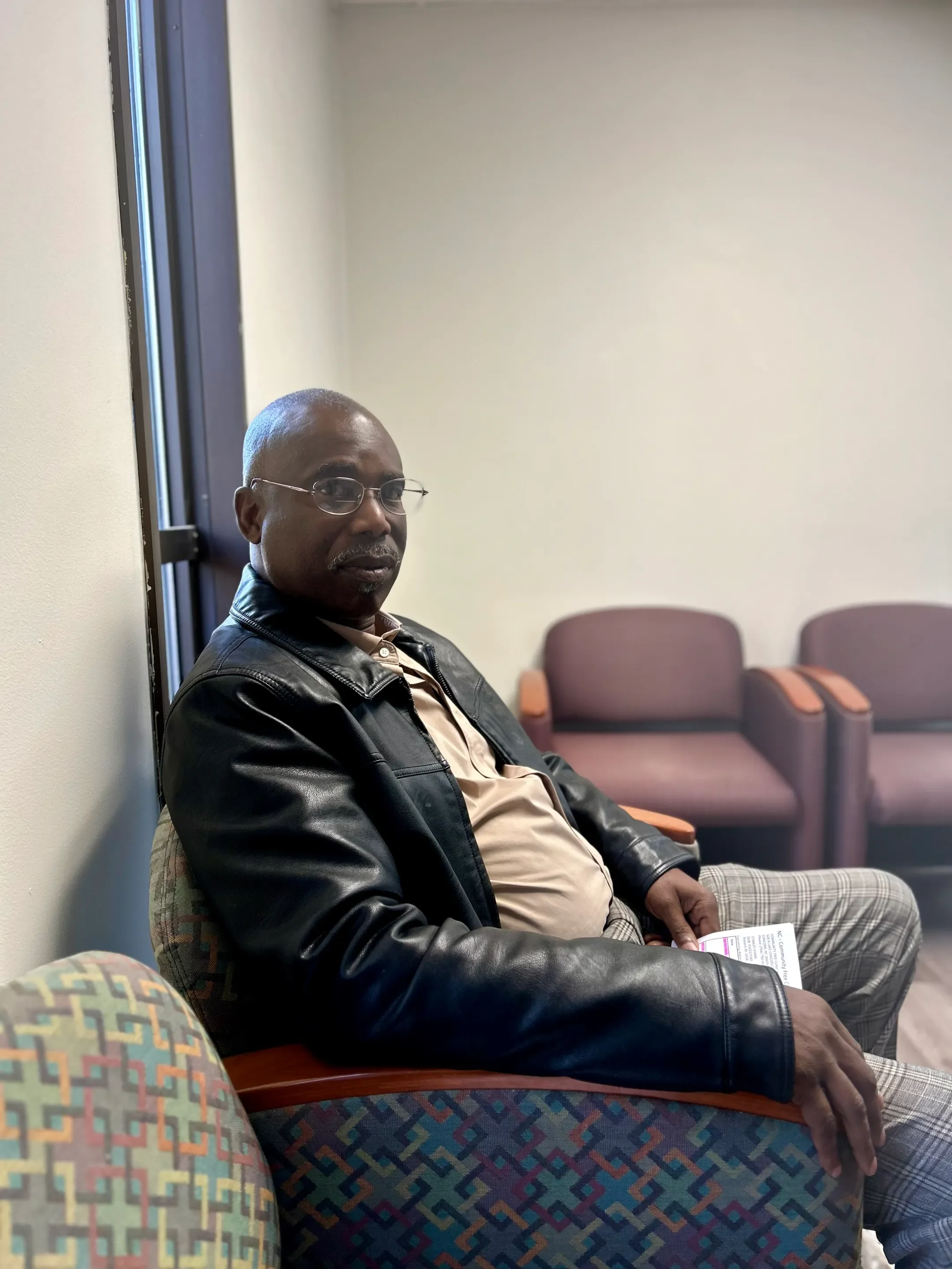 A middle-aged man with glasses and a goatee, wearing a black leather jacket, beige shirt, and plaid pants, sitting in a waiting room with brown chairs, one of which he is seated in, holding a piece of paper.