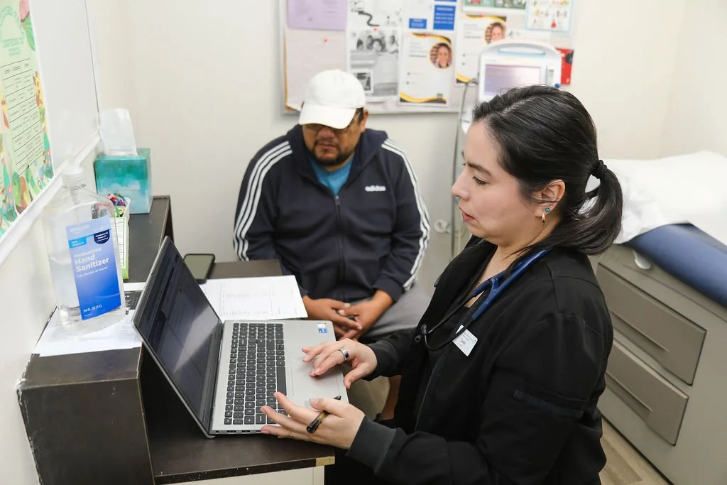 A female healthcare professional with a stethoscope around her neck is working on a laptop in a medical office. A male patient wearing a white cap and dark jacket is sitting nearby. The office has medical posters, a bed with blue bedding, and hand sanitizer on the counter.