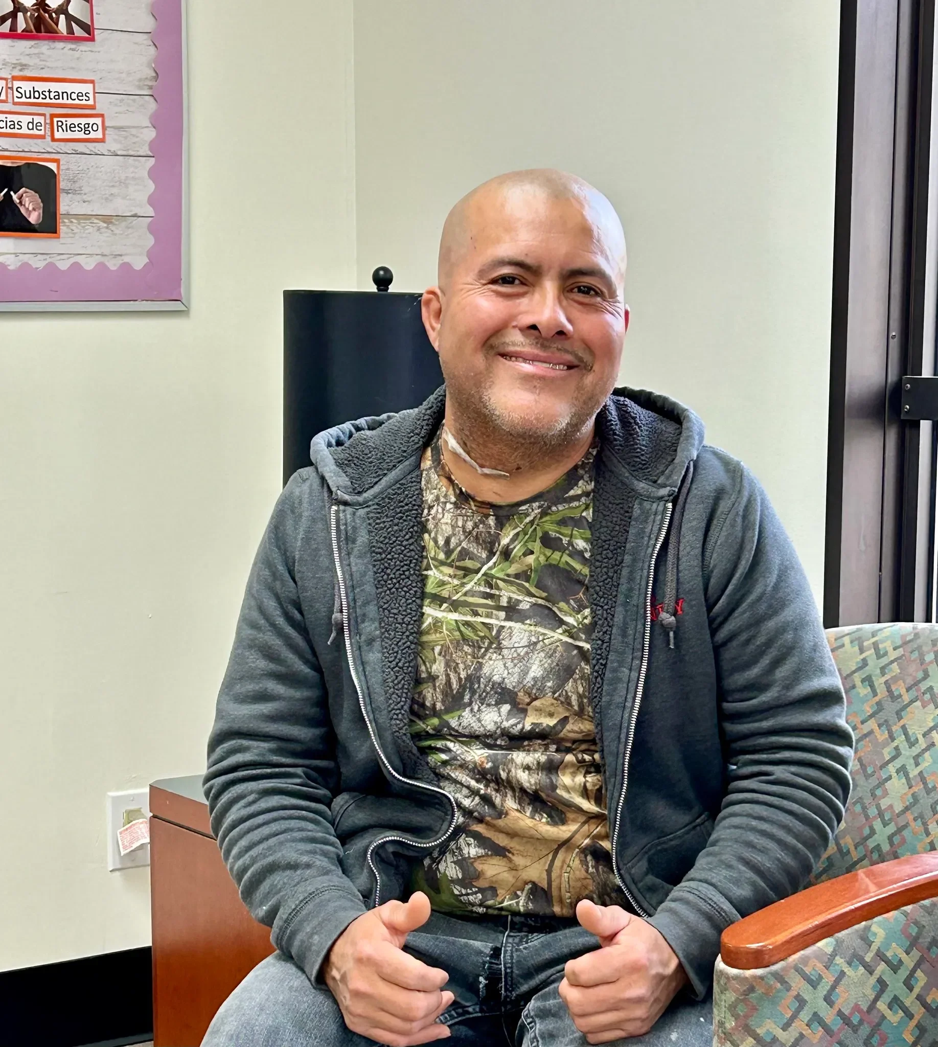 A smiling man with a bald head and beard, wearing a camouflage shirt and a dark zip-up hoodie, sitting in an office.