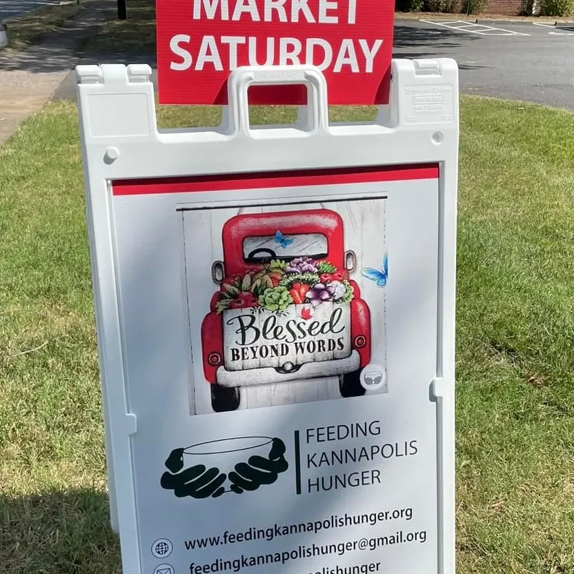 A lawn sign featuring a small red truck with a bed filled with flowers and two blue butterflies flying nearby. The sign includes text reading 'Blessed Beyond Words' on the truck bed, and additional text below: 'Feeding Kannapolis Hunger,' along with 