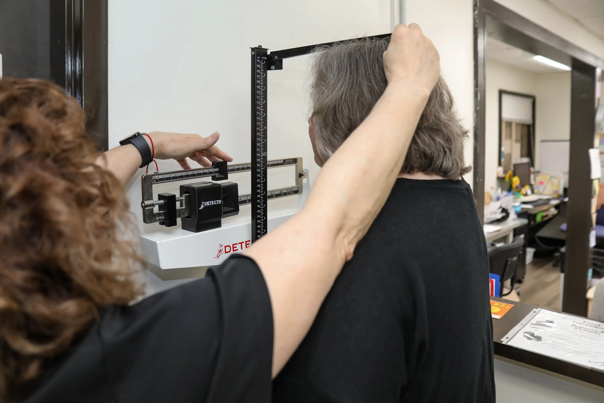 A healthcare worker measures the height of a patient using a standing height scale in an office or clinic setting.