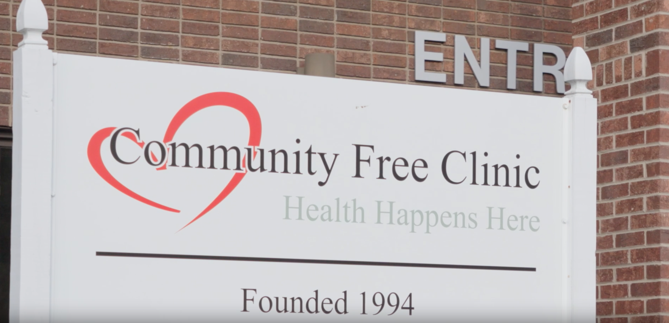 A sign for a community free clinic with a red heart graphic and text reading "Community Free Clinic," "Health Happens Here," and "Founded 1994" against a brick wall, with an "ENTRY" sign at the top.