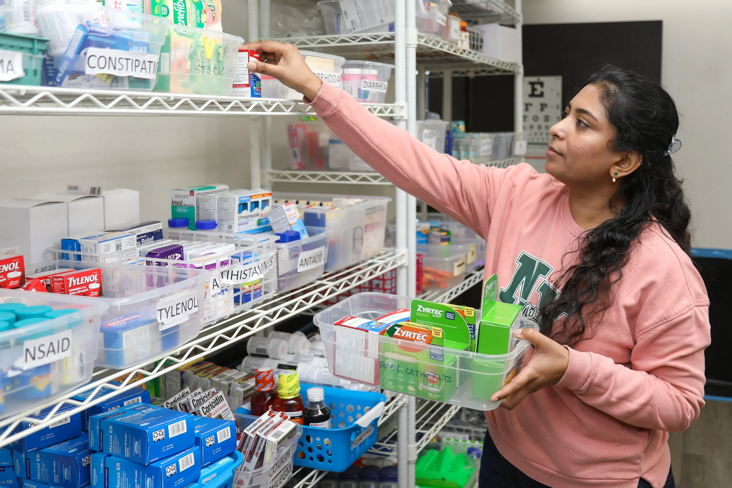 A woman shopping for medication in a pharmacy or drugstore, selecting a box from a shelf while holding a plastic container filled with medicine boxes and bottles.