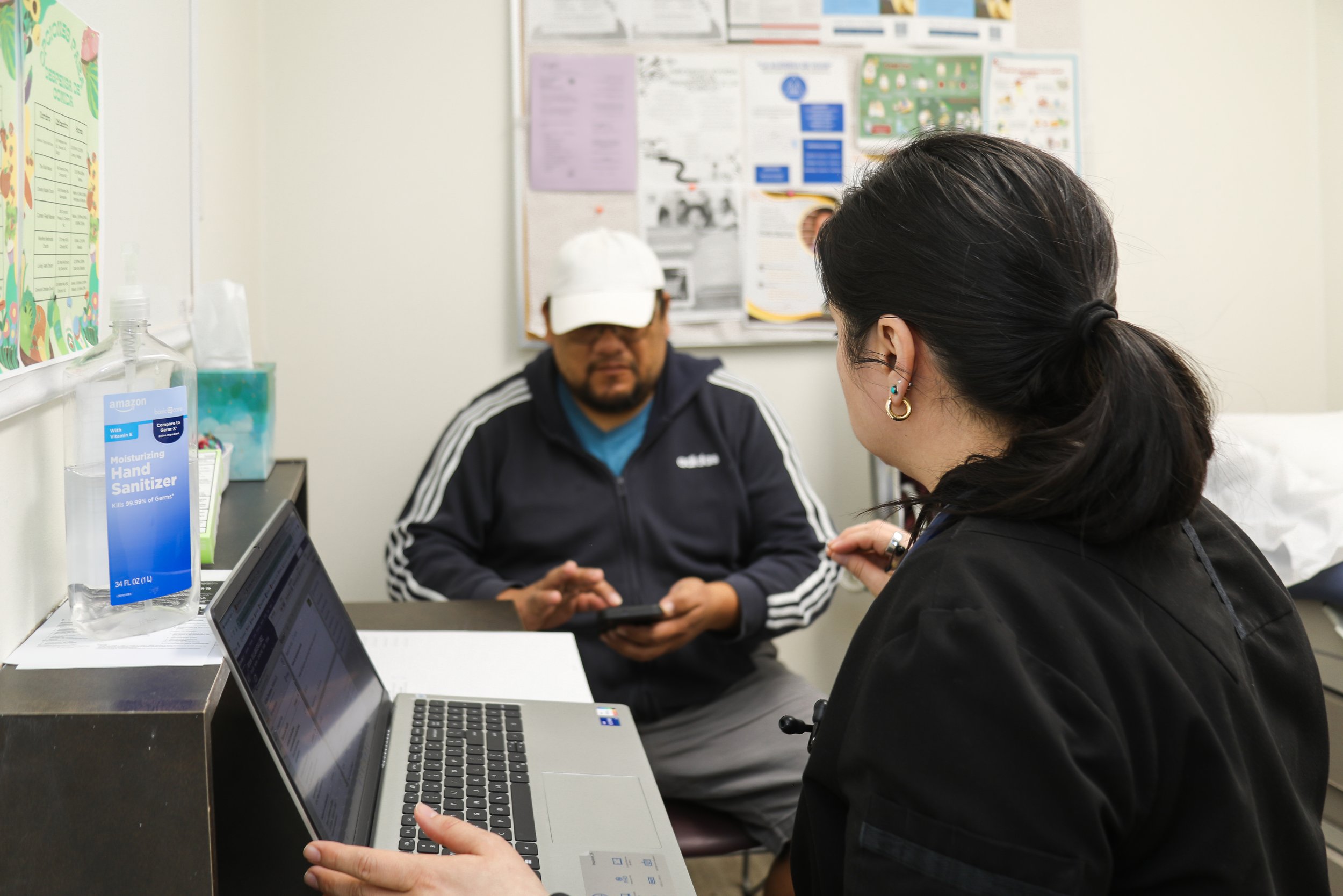 A woman and a man are sitting on either side of a desk in a medical office or clinic. The woman has dark hair tied back, multiple earrings, and is wearing a black jacket. She is using a laptop. The man is wearing a cap, glasses, a dark jacket with white stripes, and is holding a phone or electronic device. There is a bulletin board with papers and notices on the wall behind them. On the desk, there is a large bottle of hand sanitizer and some papers.