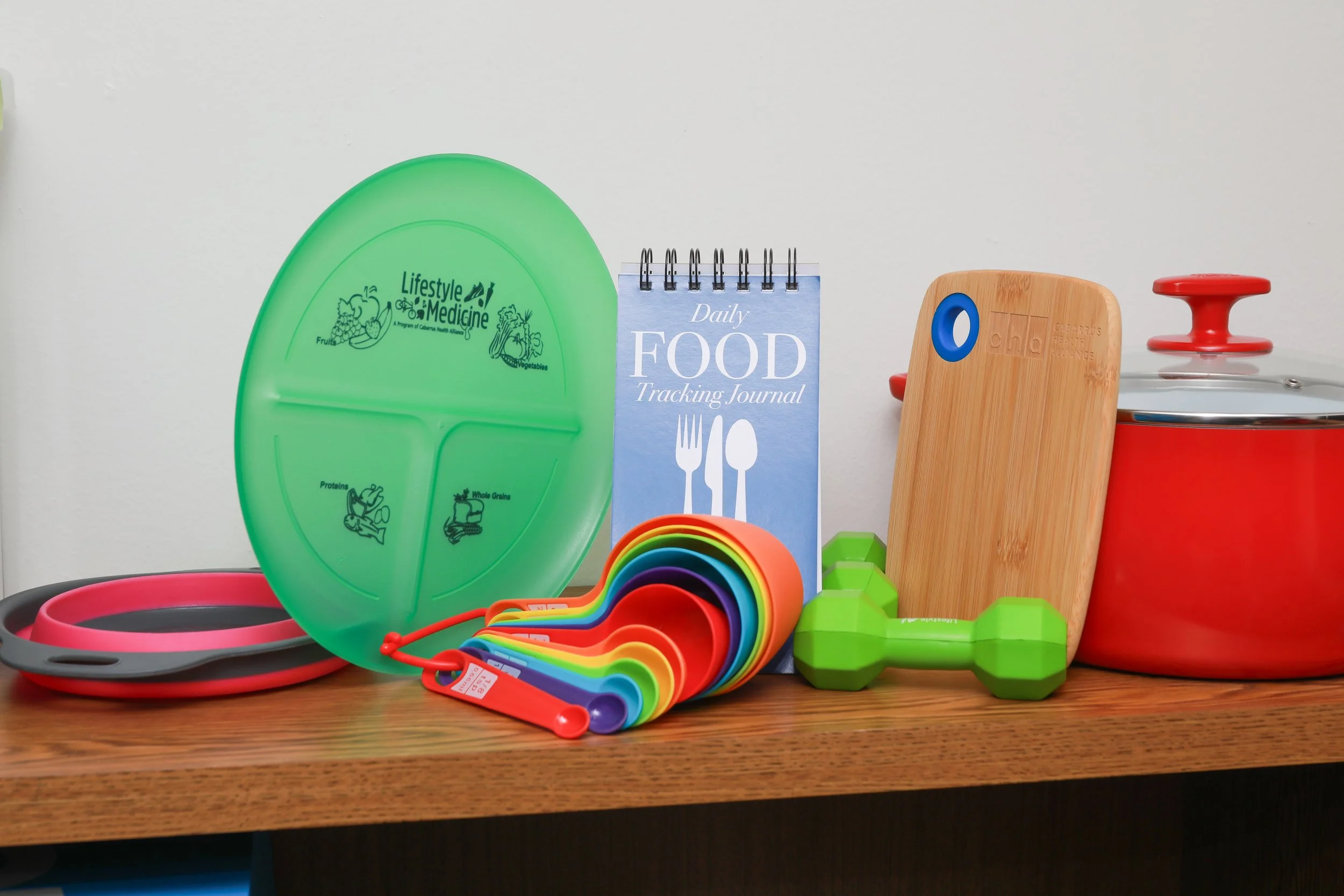 A colorful collection of kitchen and fitness items on a wooden table, including a green divided plate, rainbow measuring cups, a wooden cutting board, green weights, and a red pot with a lid.