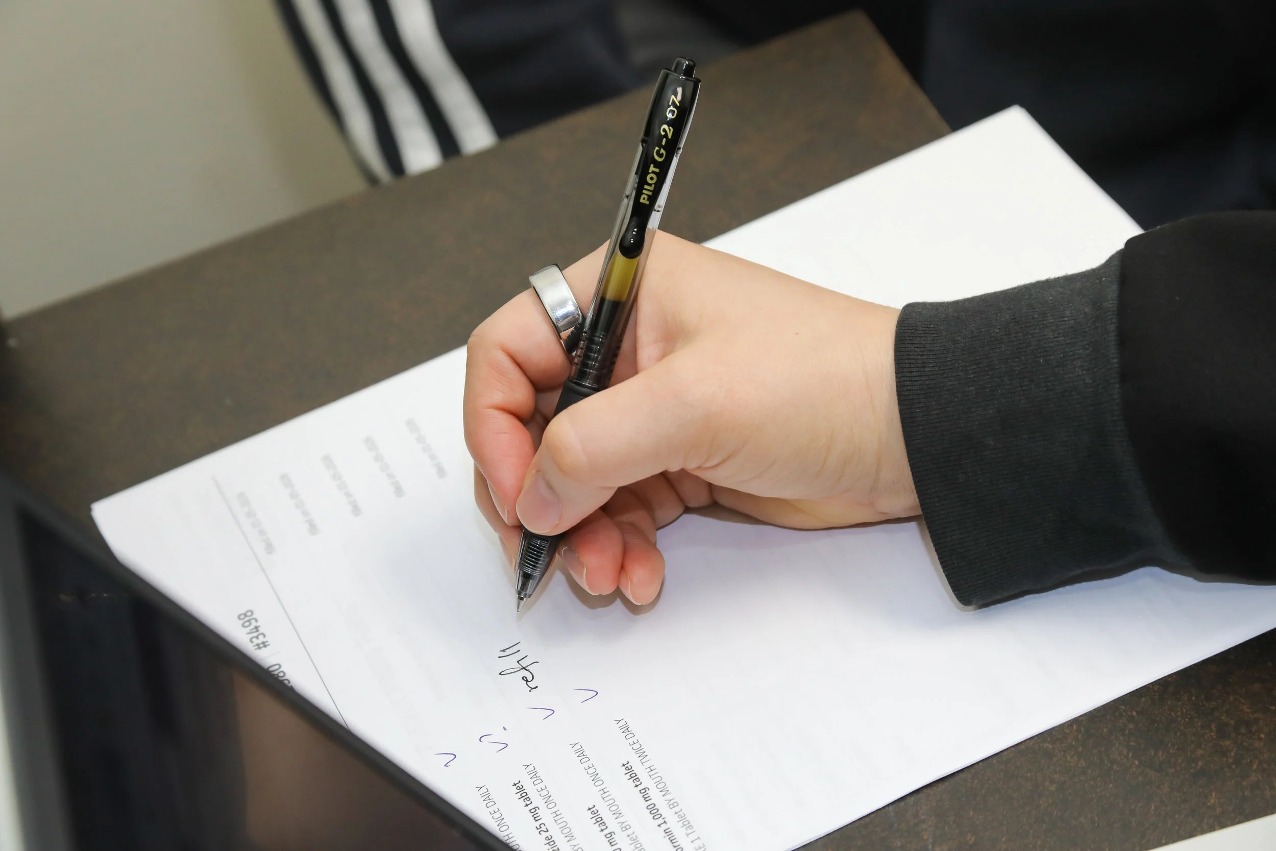 A person wearing a black hoodie with a silver ring on their finger is writing on a white sheet of paper with a black pen. The paper is on a dark brown surface, and some text is visible on the sheet.