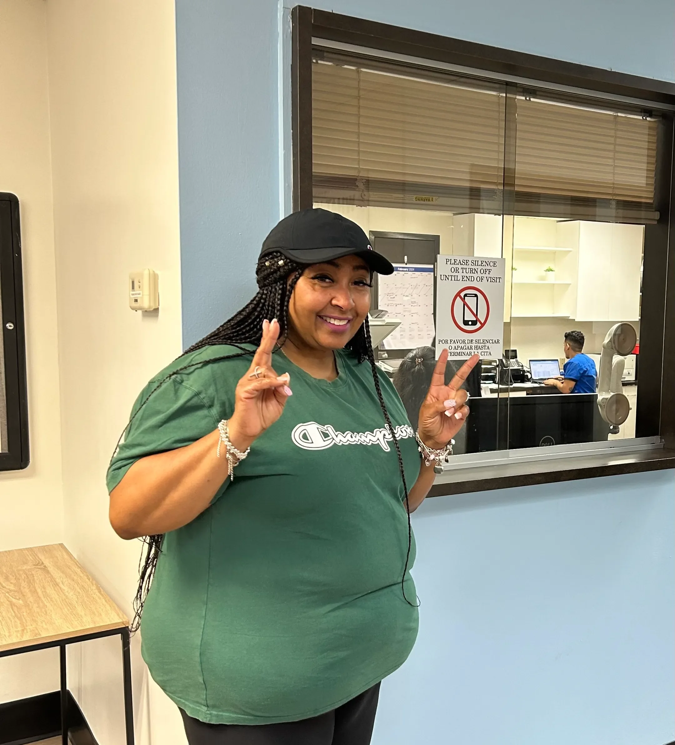Smiling woman with braided hair and a black cap making peace signs at a medical or office reception window.