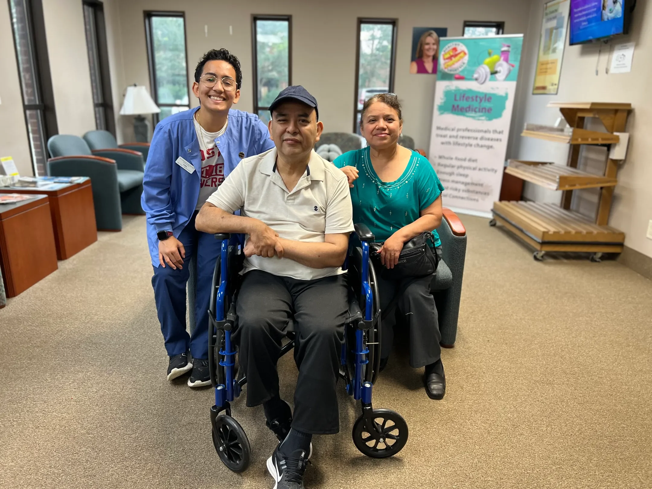 Three people posing in a the Community Free Clinic Cabarrus's waiting area. Two women and one man in a wheelchair. There are chairs, a poster about lifestyle medicine, and windows in the background.