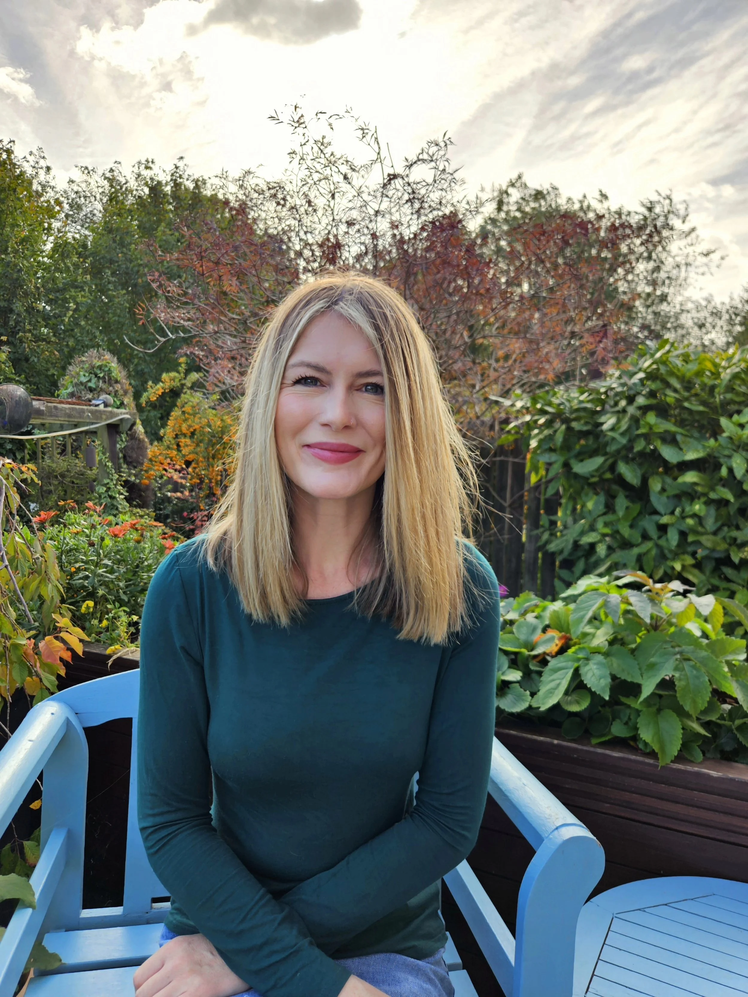 A woman with blonde hair and a green long-sleeve shirt sitting on a blue bench in a garden with colorful plants and trees, under a partly cloudy sky.