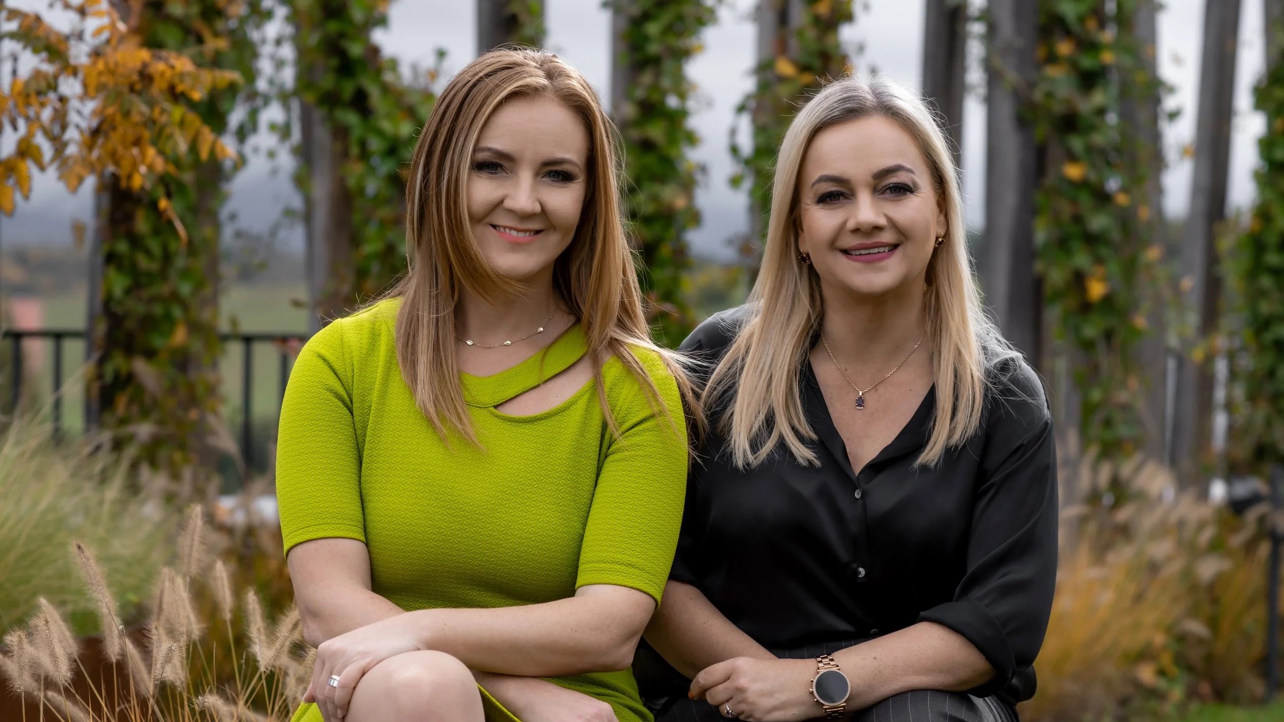 Two women sitting outdoors in front of a vine-covered structure, smiling at the camera.