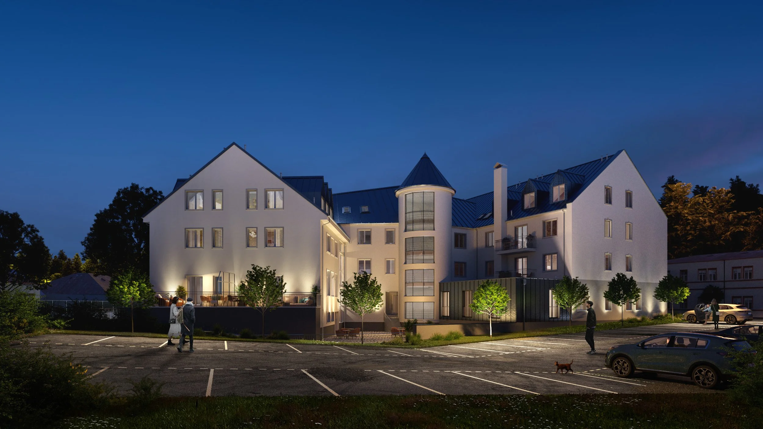 A modern multi-story residential building illuminated at night, with trees and parked cars in the foreground, and people walking in a parking lot.