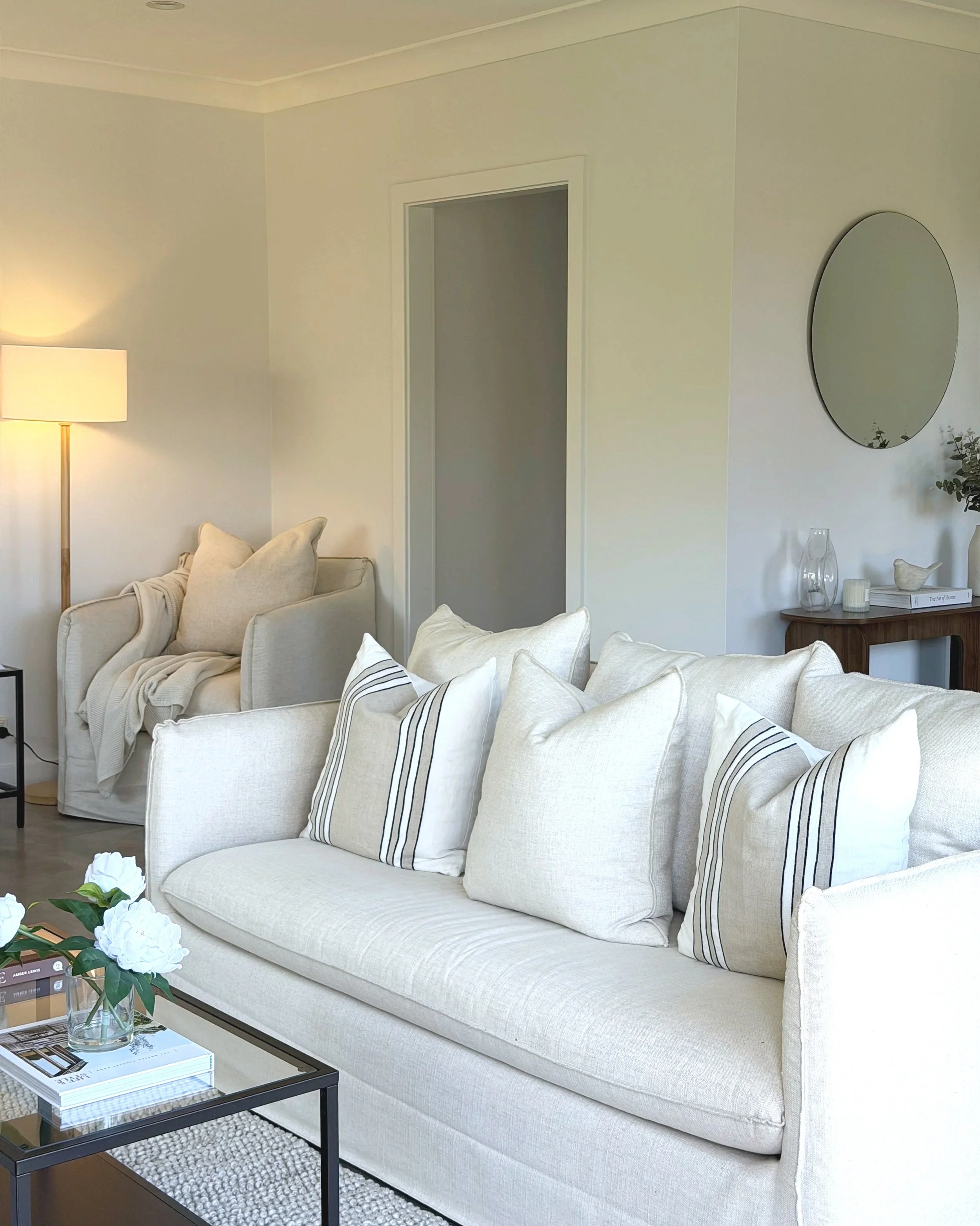 Living room with beige sofa, armchair, and various decorative pillows, pillows have beige and white striped covers. Coffee table with magazines and a flower vase. Background includes a side table with vases and decorative items, a round mirror, and a