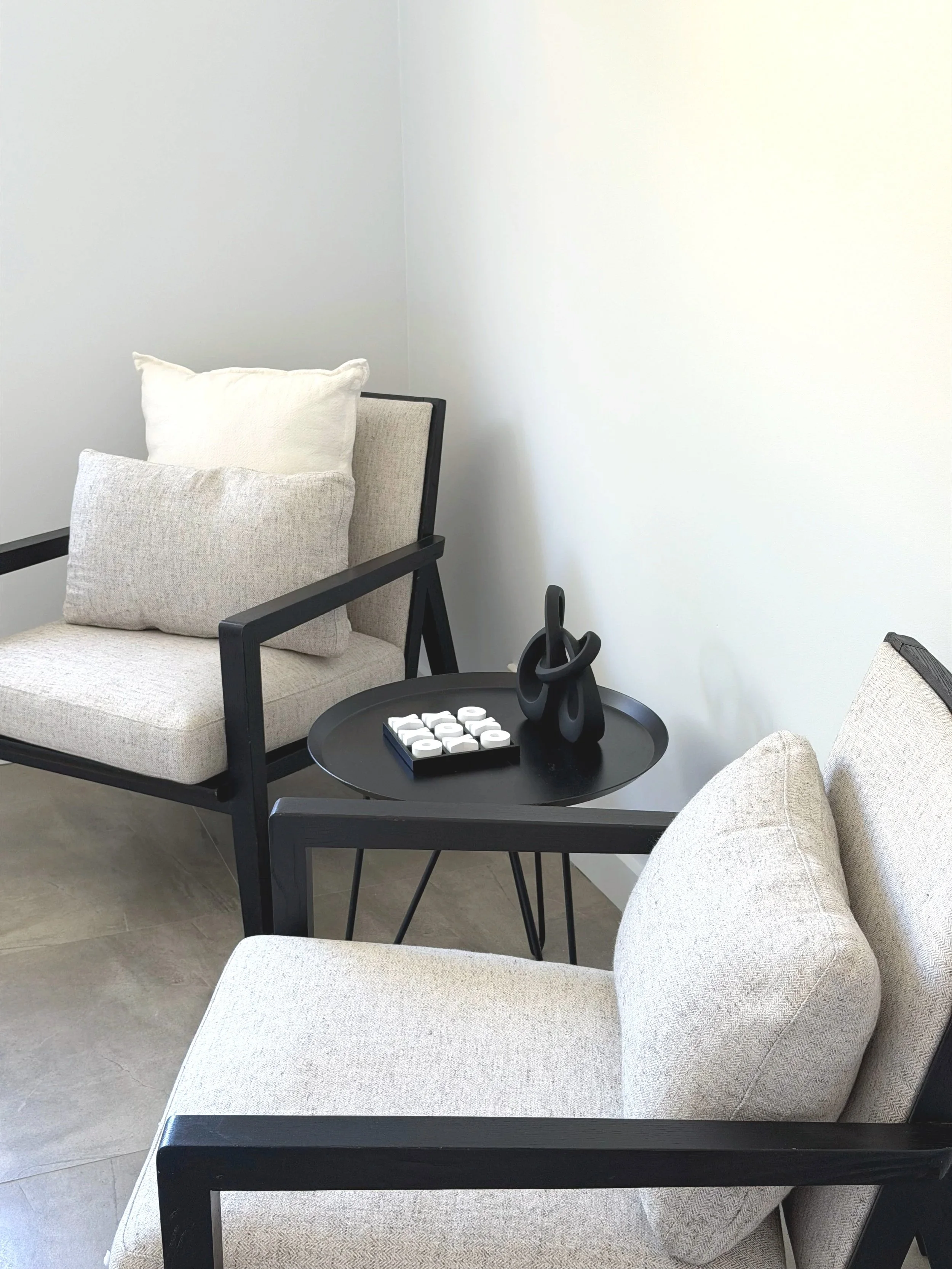 A modern living room corner with two beige upholstered armchairs with black frames, positioned opposite each other. A small black round table between them holds decorative black sculptures and a tray with white candles.
