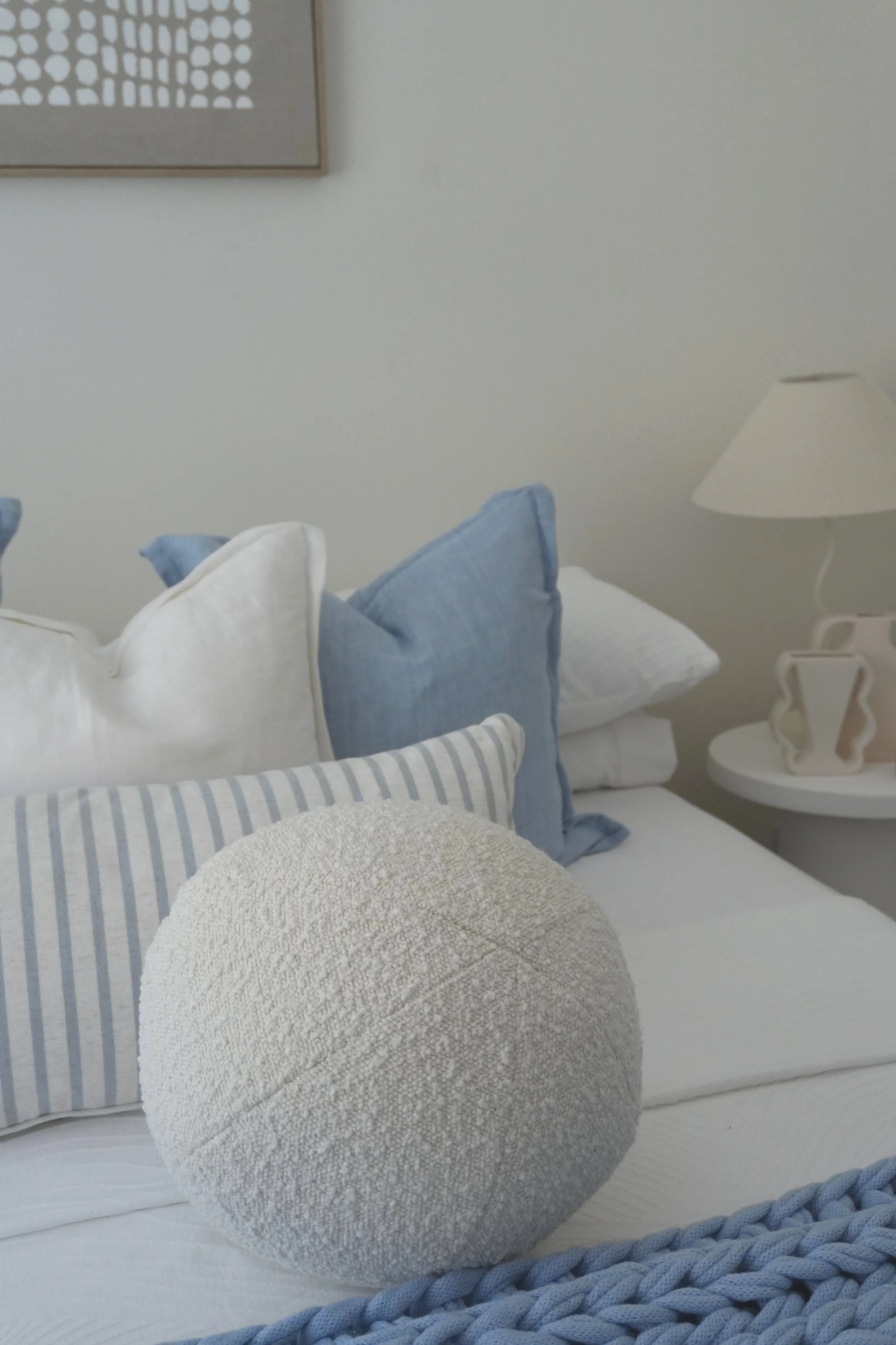 A neatly made bed with white, blue, and striped pillows, a textured round pillow in the foreground, and a bedside table with a lamp and decorative object in a bedroom with white walls.