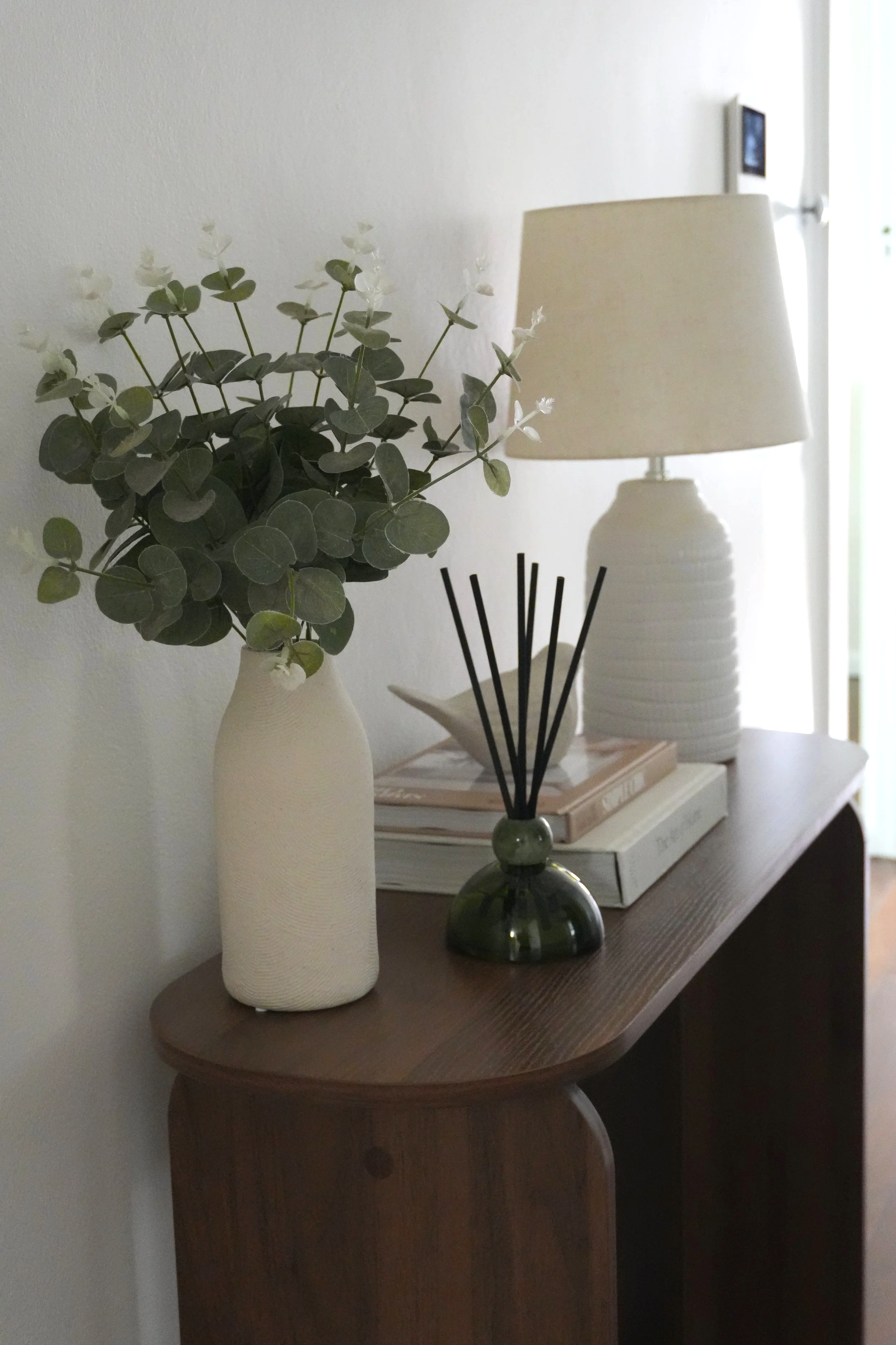 Decorative wooden side table with a white vase containing eucalyptus branches, a small black reed diffuser, a stack of books, and a white textured table lamp in a room with natural light.