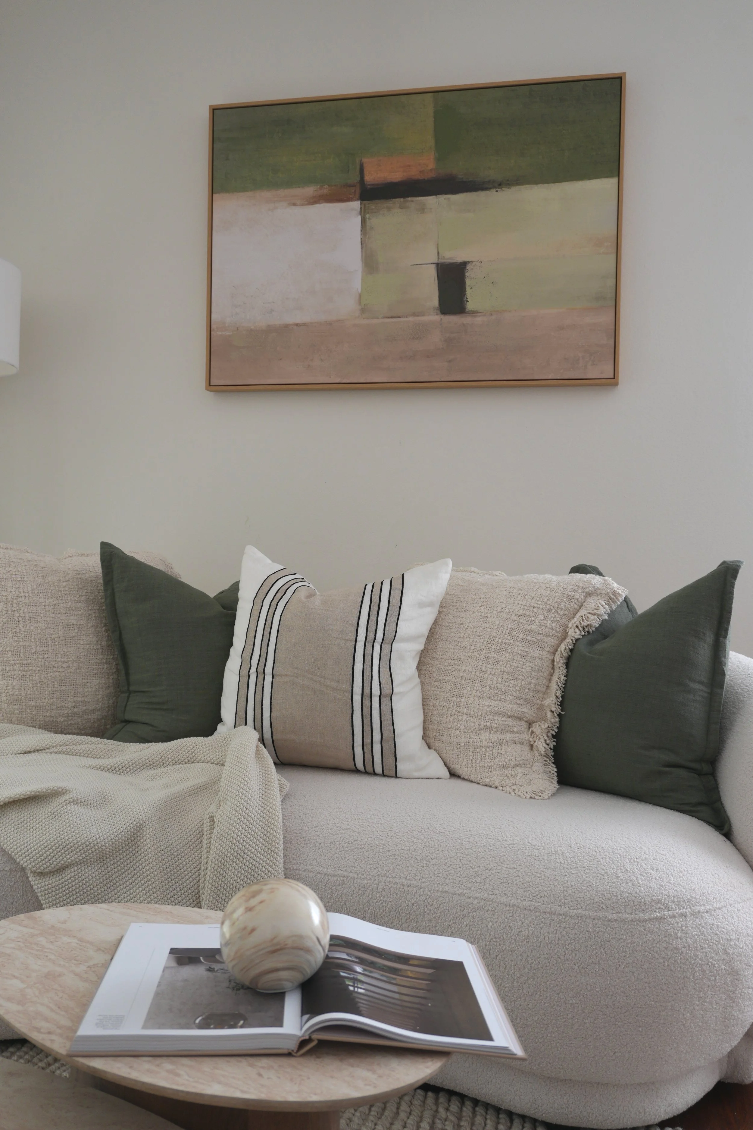 Living room with a white textured sofa, beige and dark green pillows, a round marble coffee table with an open magazine and a decorative stone, a beige and white throw blanket, wall art with abstract green, beige, and black shapes.