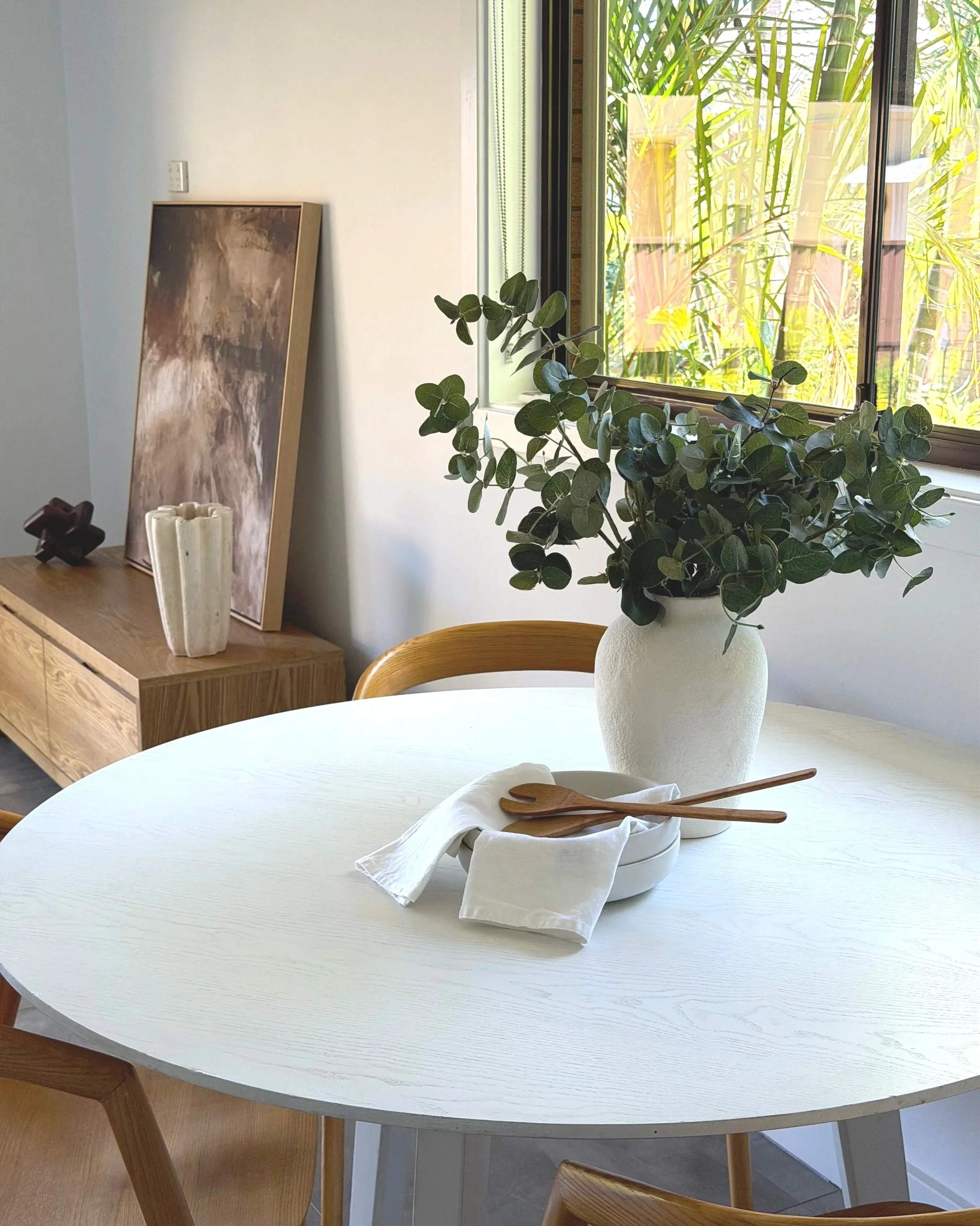 A white round dining table with a potted green plant, wooden utensils, and white cloth napkins. In the background, there is a wooden sideboard with decorative items and artwork leaning against the wall. An open window shows greenery outside.
