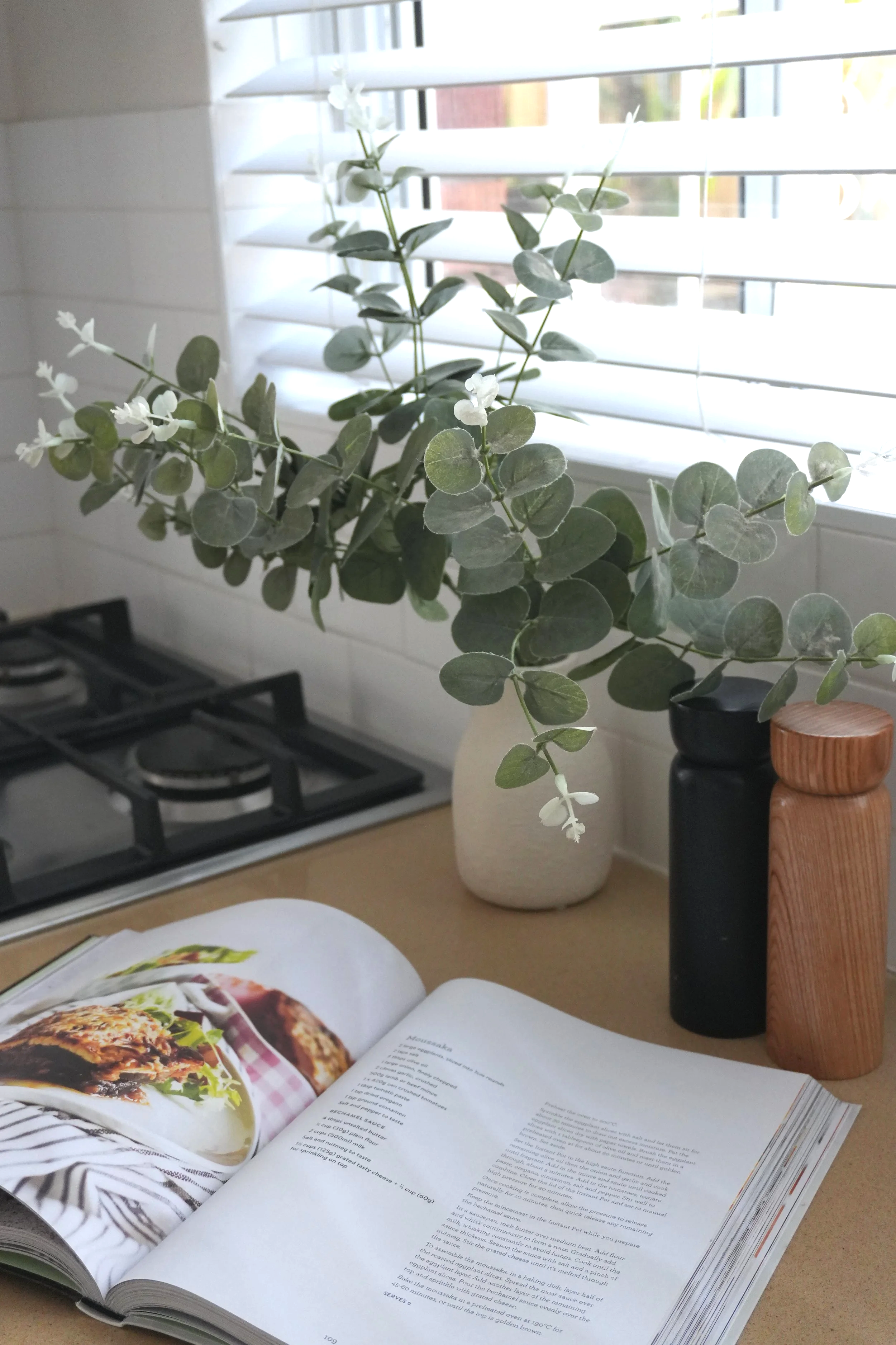 A kitchen countertop with a vase holding green foliage, an open cookbook showing a dish, a black spice jar, and a wooden pepper mill near a window with blinds.