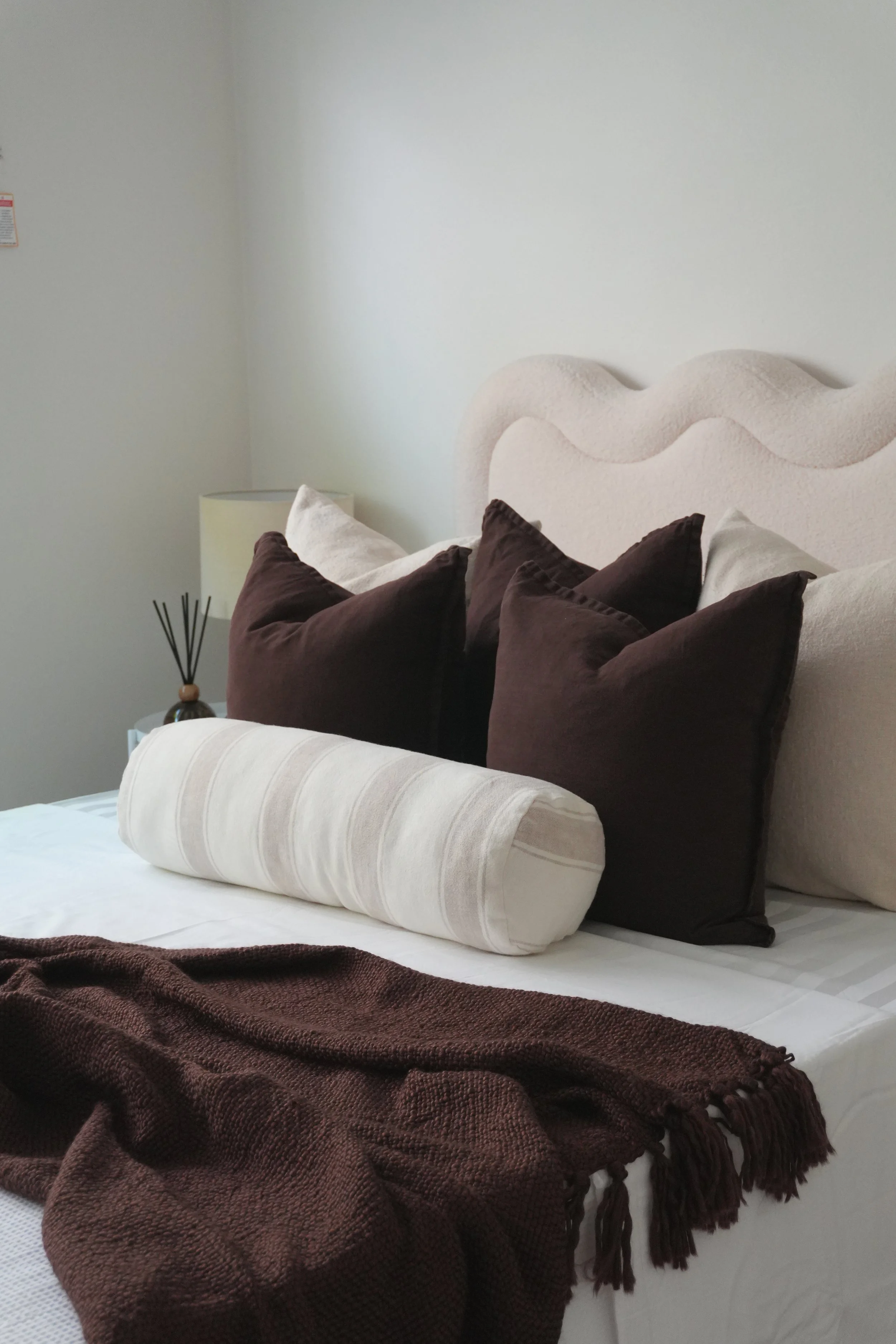 A neatly made bed with dark brown pillows, a beige bolster pillow, a textured brown throw blanket, and a beige headboard against a white wall, with a small nightstand and lamp visible on the side.