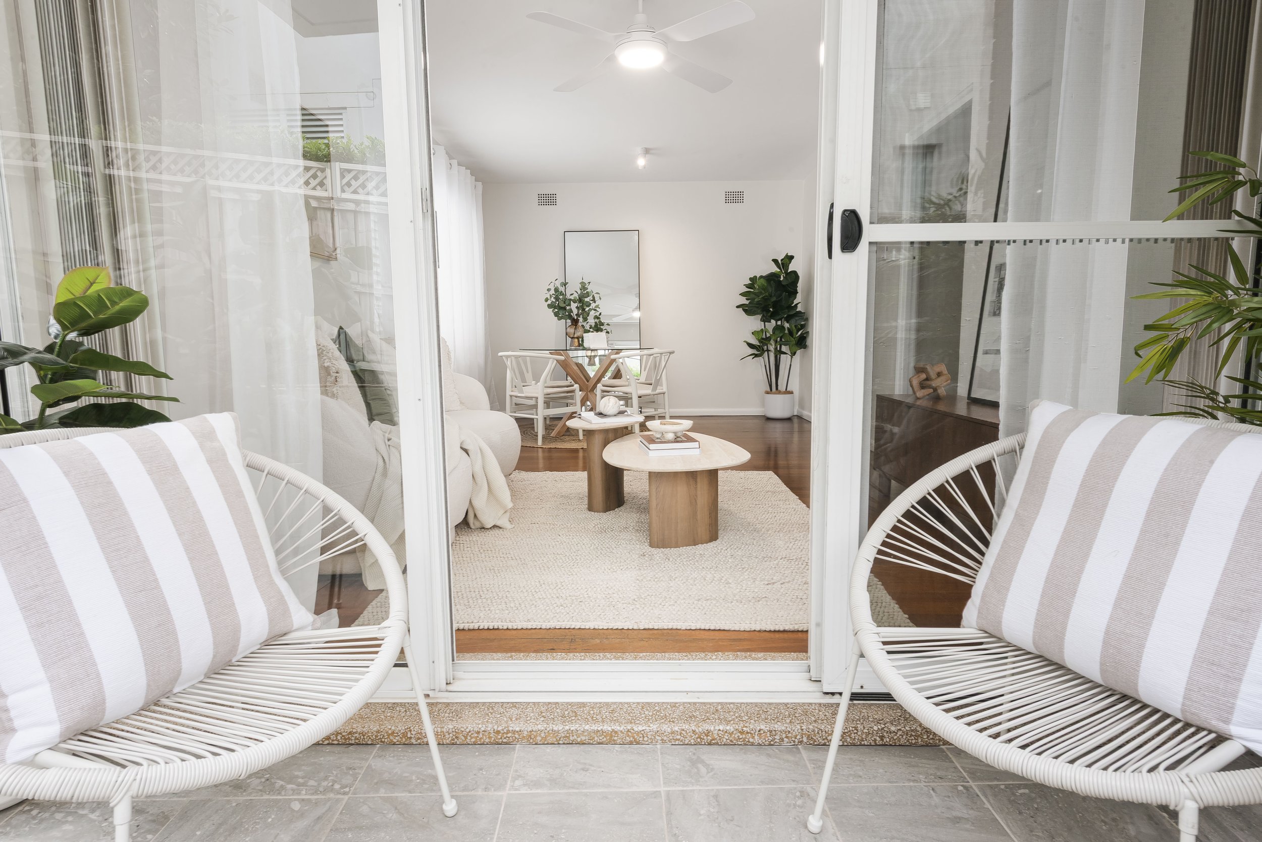 View through sliding glass doors into a bright, modern living room with white furniture, wooden coffee table, and indoor plants.