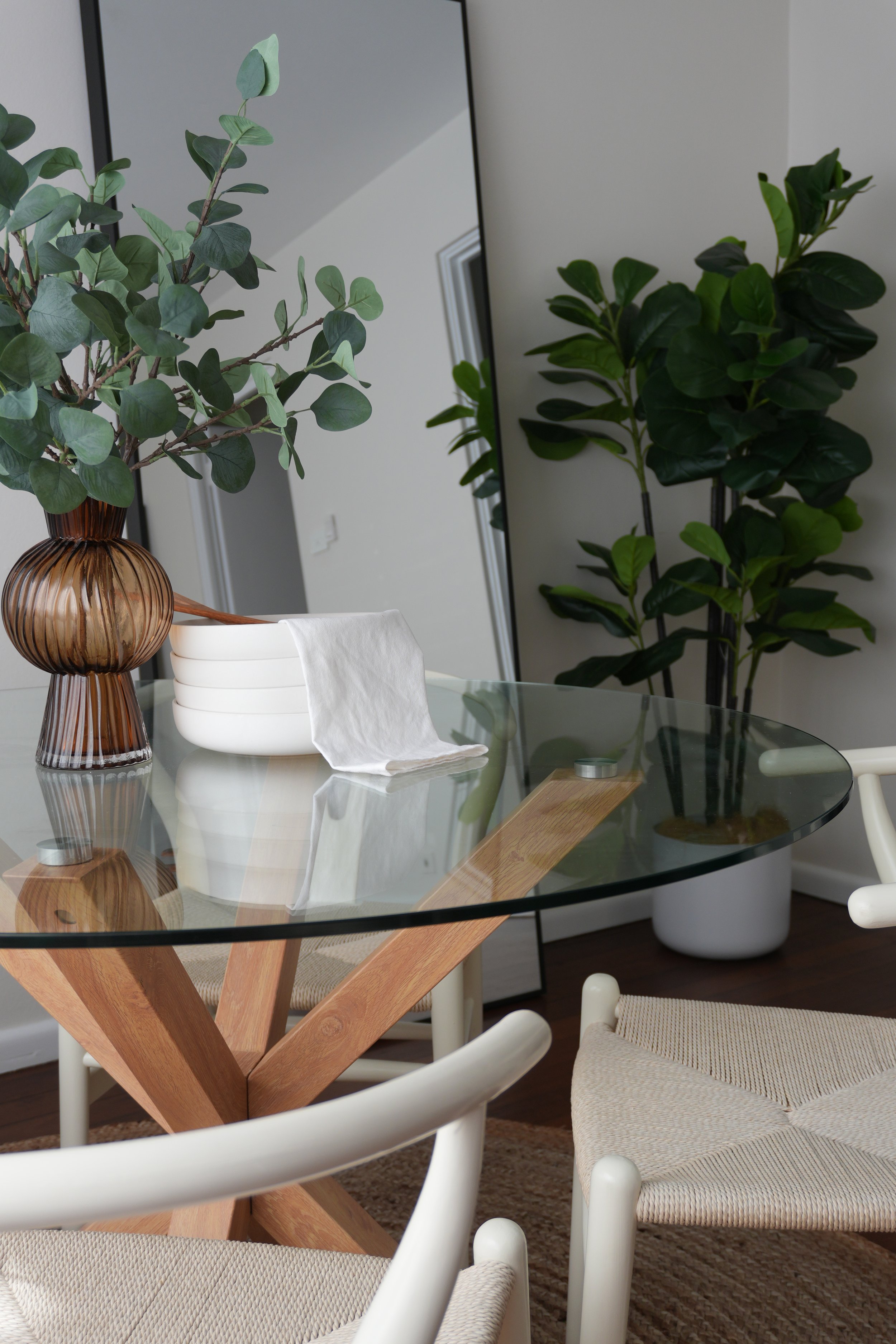 Decorative glass table with a wooden cross-shaped base, surrounded by white woven chairs, with a large potted plant and a vase with eucalyptus branches on the table.