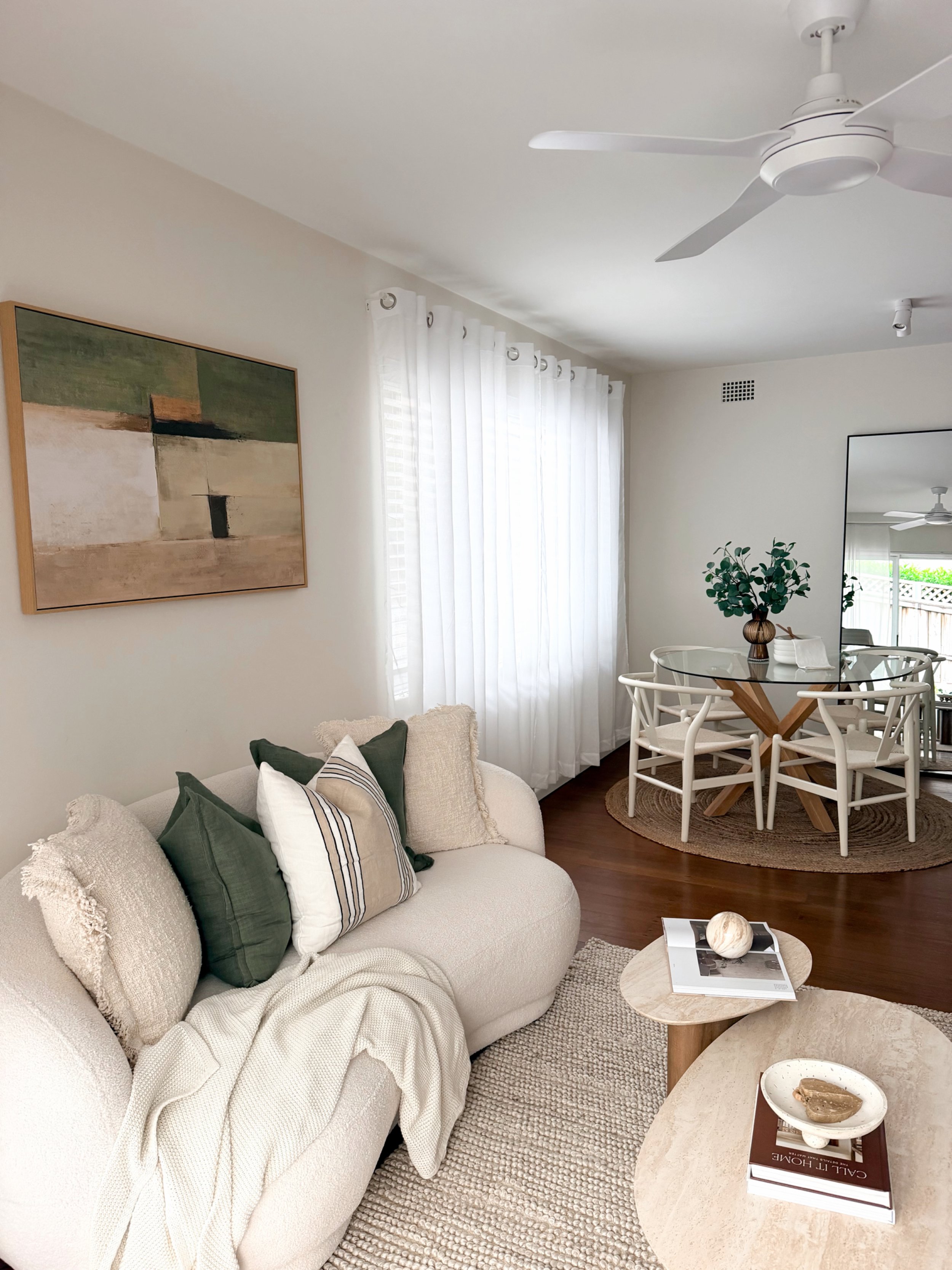A bright, modern living room with white walls, a cream-colored sofa with green and patterned pillows, a beige textured rug, and a wooden coffee table with decorative stones and books. In the background, there is a round dining table with four white chairs, a large mirror, and a potted plant on the table. White curtains cover the window, and a ceiling fan is installed on the ceiling.