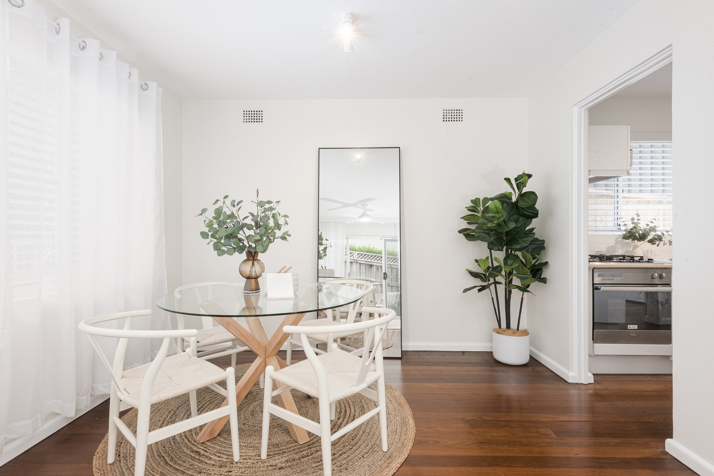 A dining area with a glass-top table surrounded by white chairs on a round woven rug. A large mirror leans against the wall, with a vase of green leaves on the table. A tall potted plant is next to an open doorway leading to a kitchen.
