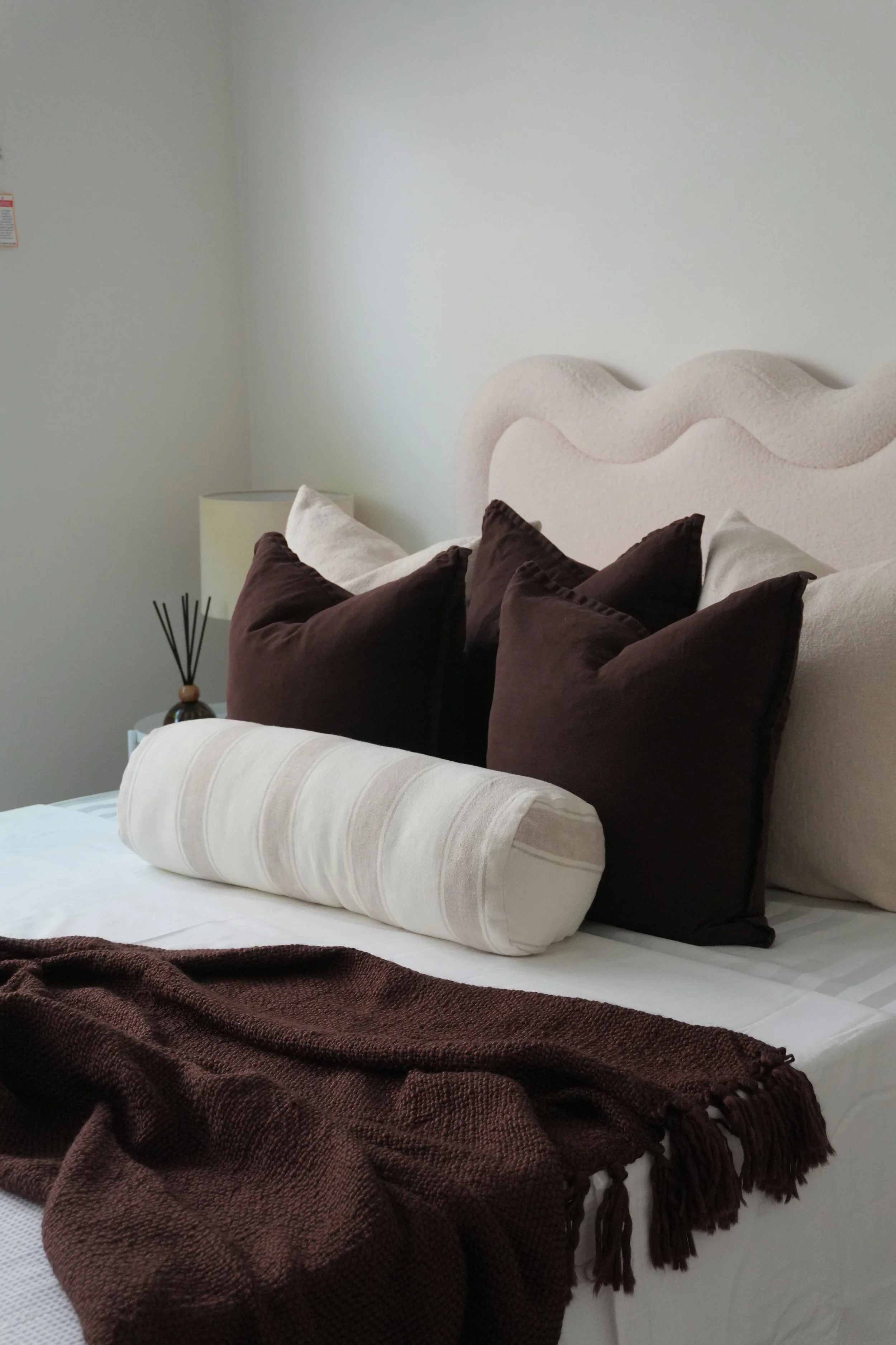 A neatly made bed with a beige, wave-patterned headboard, topped with multiple pillows including dark brown, light beige, and a beige cylindrical bolster, with a brown throw blanket at the foot and a bedside lamp with a reed diffuser on a nightstand.