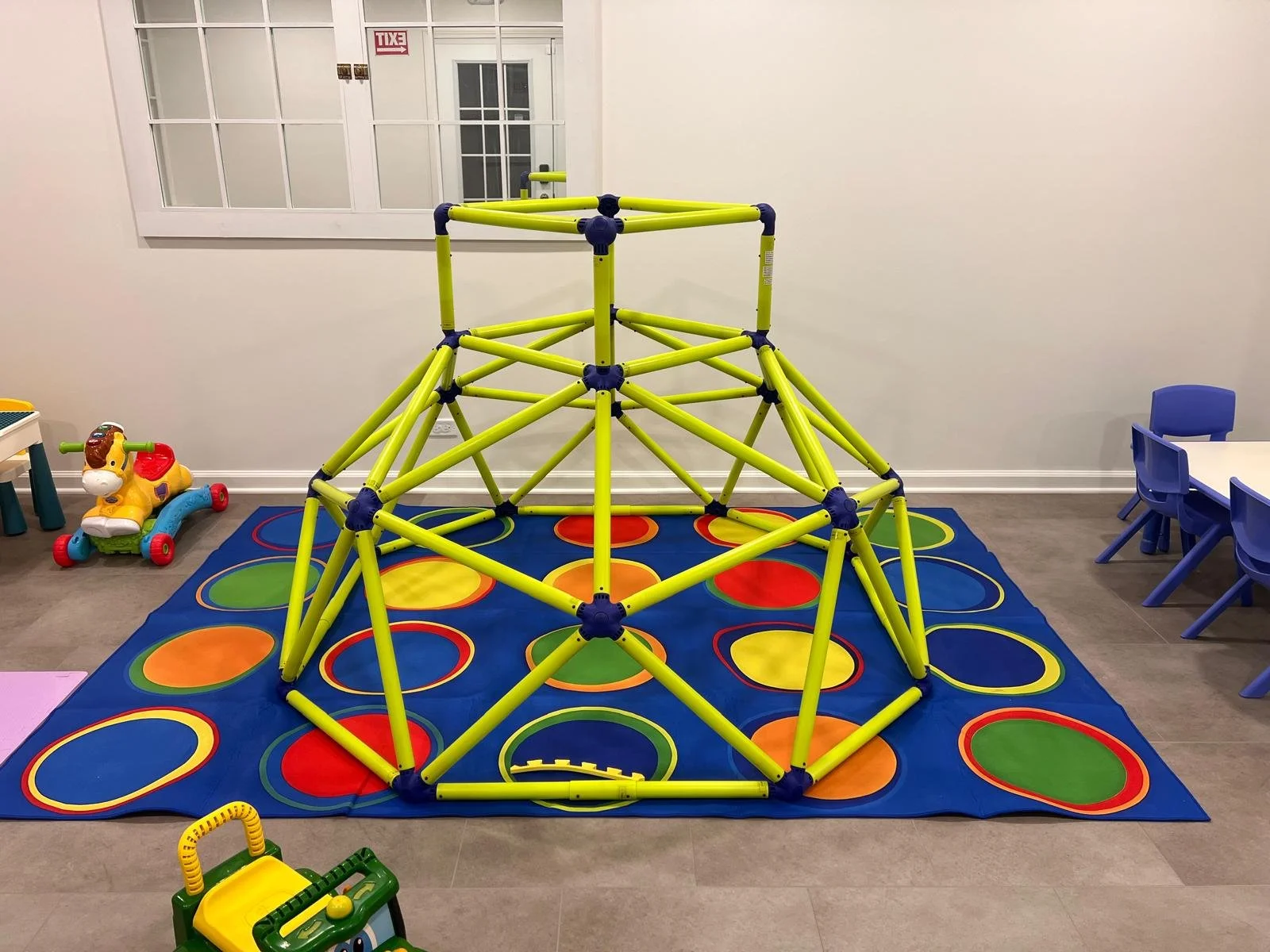 Indoor children's play area with a yellow climbing structure on a blue and multicolored circular patterned mat, a toy dog on wheels to the left, and small blue tables and chairs to the right. There is a white wall with a window and a door in the back