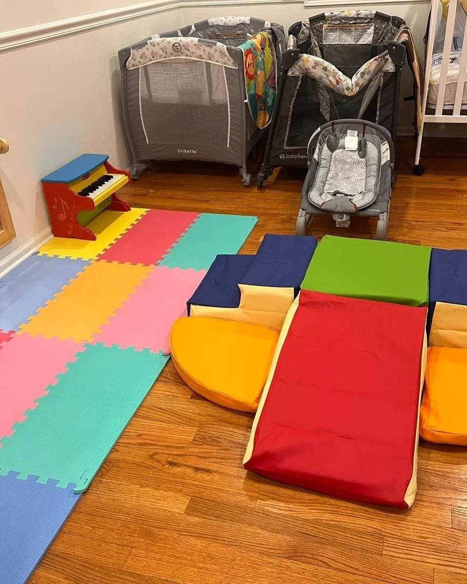 A playroom with colorful foam puzzle mats on the wooden floor, small toys, a mirror, cribs, a toy piano, a high chair, and various baby gear including strollers and a boxed humidifier.