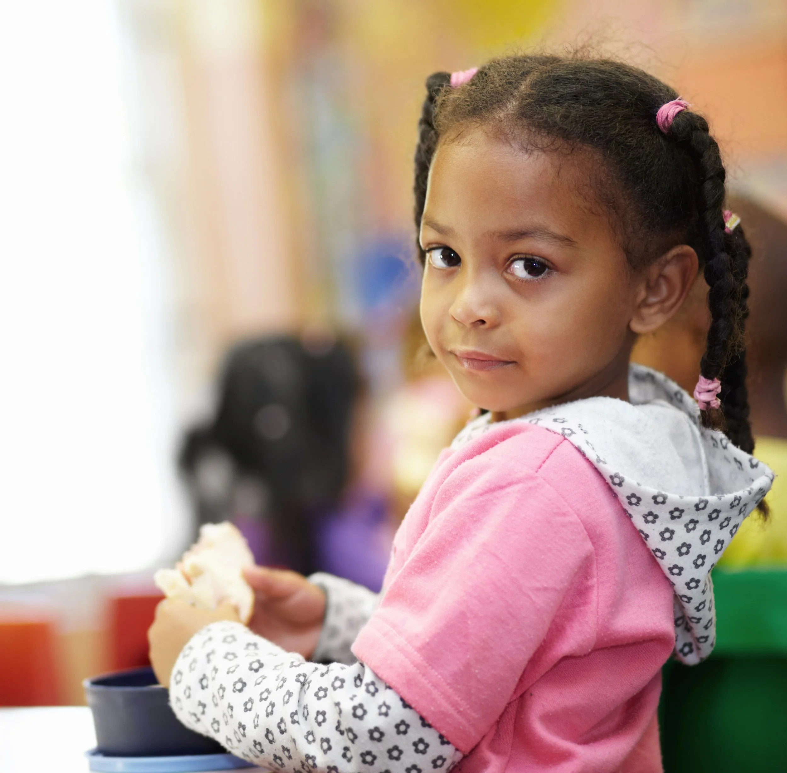 A young girl with braided hair and pink hair ties, wearing a pink and white hoodie, looks at the camera while holding a sandwich in her hands.