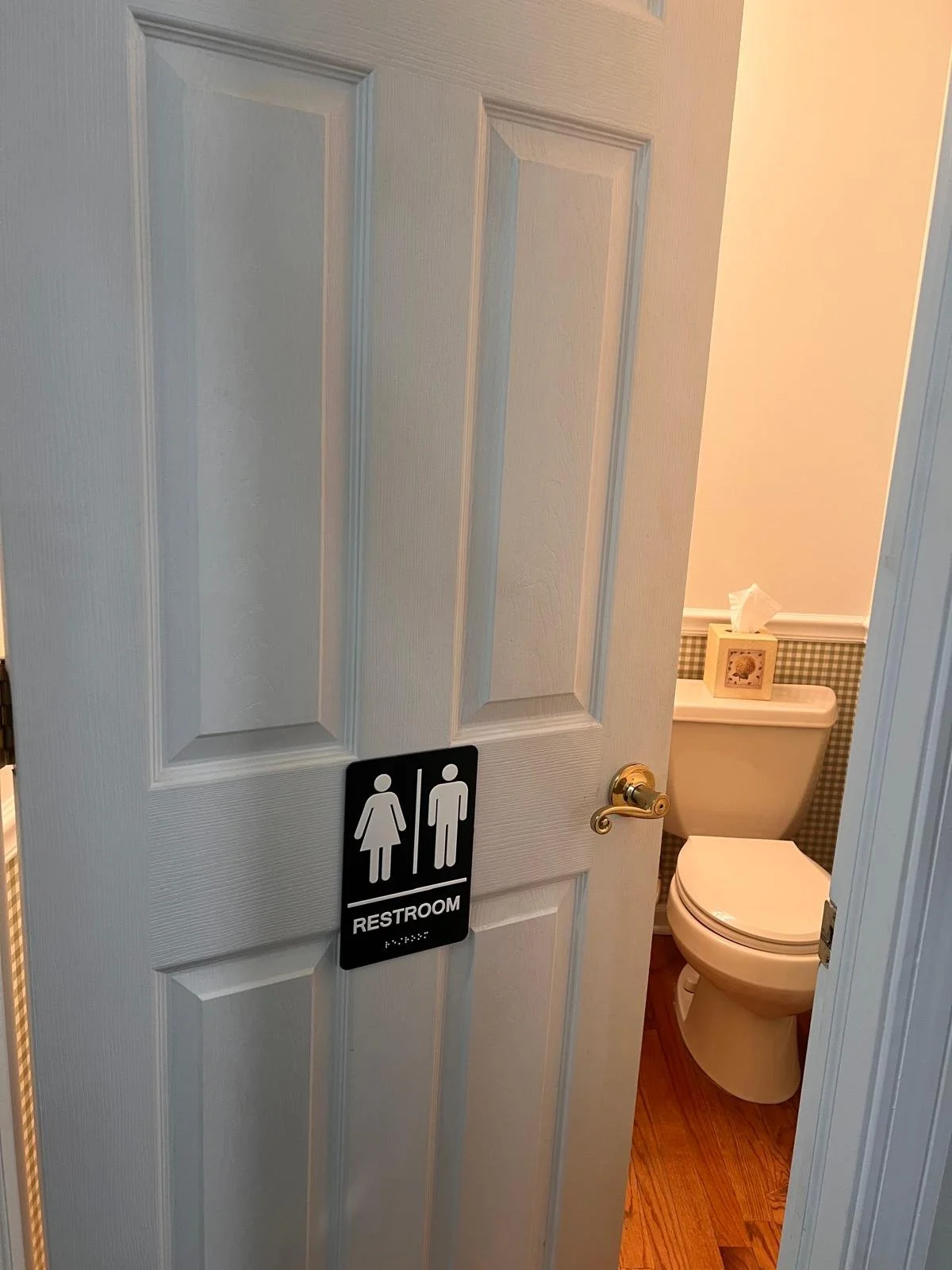 A partially open white bathroom door with a black and white sign that says "Restroom" and features icons of a woman and a man. Inside, there is a white toilet with a box of tissues and a framed picture on top, and a small section of wallpaper with a 