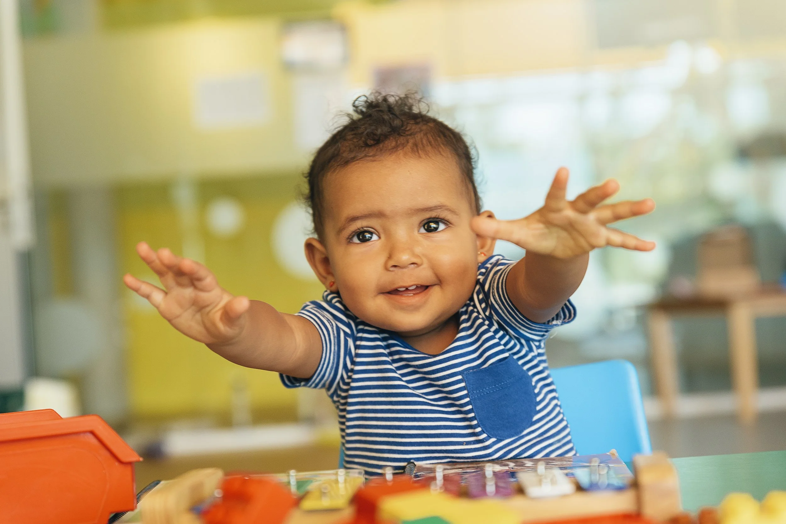 A young child with curly hair and brown skin smiling and reaching out with both hands, sitting at a table in a colorful room.