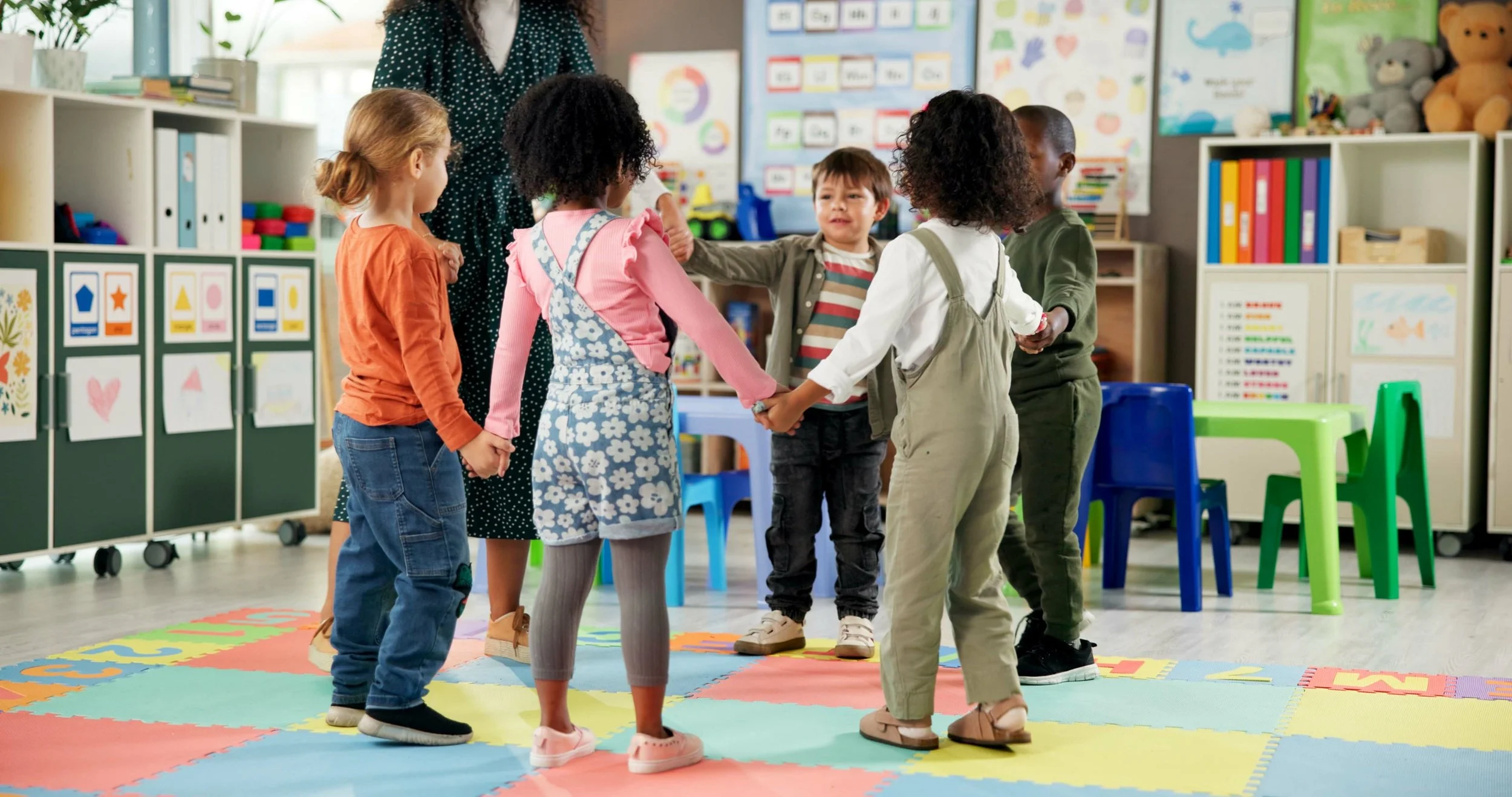 Six children holding hands in a circle in a colorful classroom, with a teacher standing nearby and educational posters on the walls.