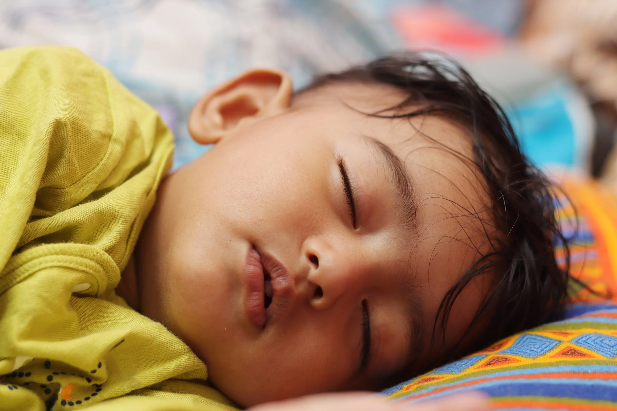 Close-up of a young child sleeping peacefully on a colorful pillow, wearing a yellow shirt.