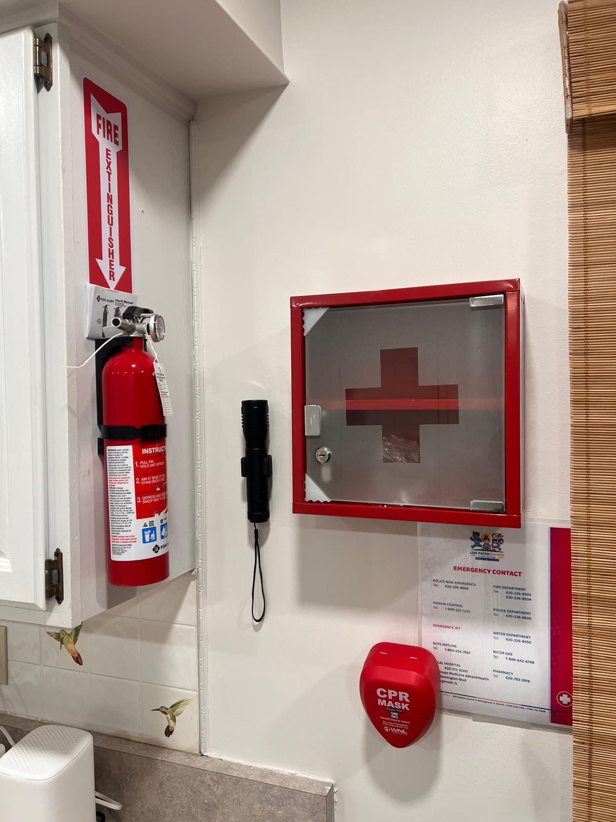 Fire extinguisher, red first aid kit labeled 'CPR Mask,' emergency contact information printed on a sheet of paper, and a red cross sign on an enclosed cabinet, all mounted on a white wall in a public safety area.