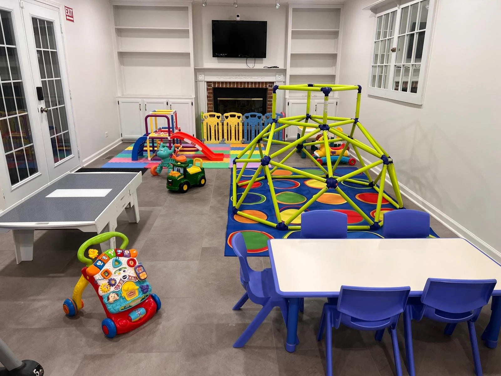 Indoor playroom with colorful play structures, toys, small chairs, and a table. It has a TV mounted above a fireplace on the back wall, with a rainbow-colored hopscotch mat on the floor and a multicolored climbing frame. There are windows and a set o