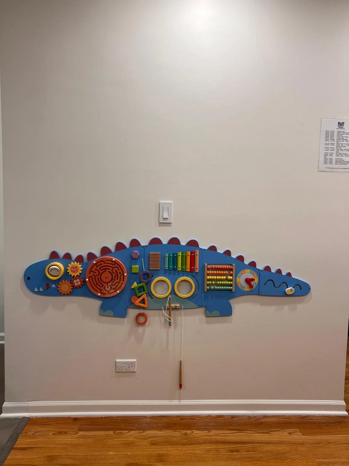 Colorful children's wall toy shaped like a dinosaur with various interactive elements such as gears, rings, and abacus on a beige wall in a playroom.