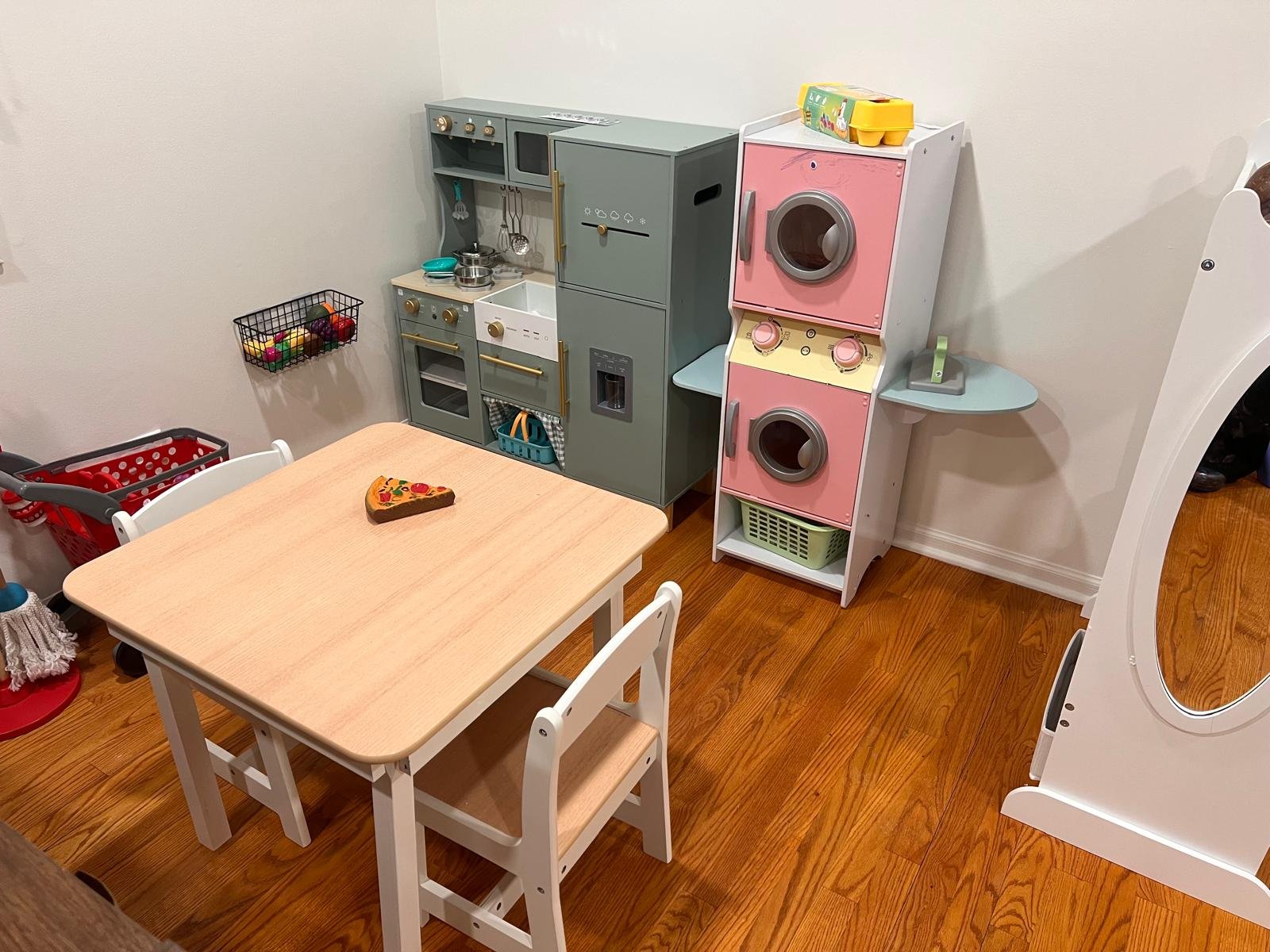 A children's play kitchen set with a stove, oven, and microwave, a pink toy washing machine, a small table with a pizza-shaped toy, and other small accessories in a playroom with wooden flooring.