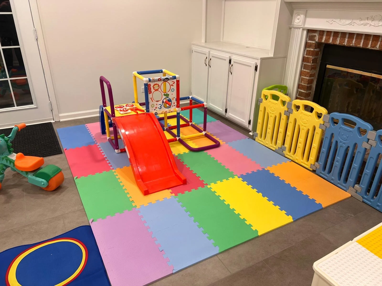 Indoor children's play area with colorful foam mats, a small slide, a climbing structure, and plastic safety gates.
