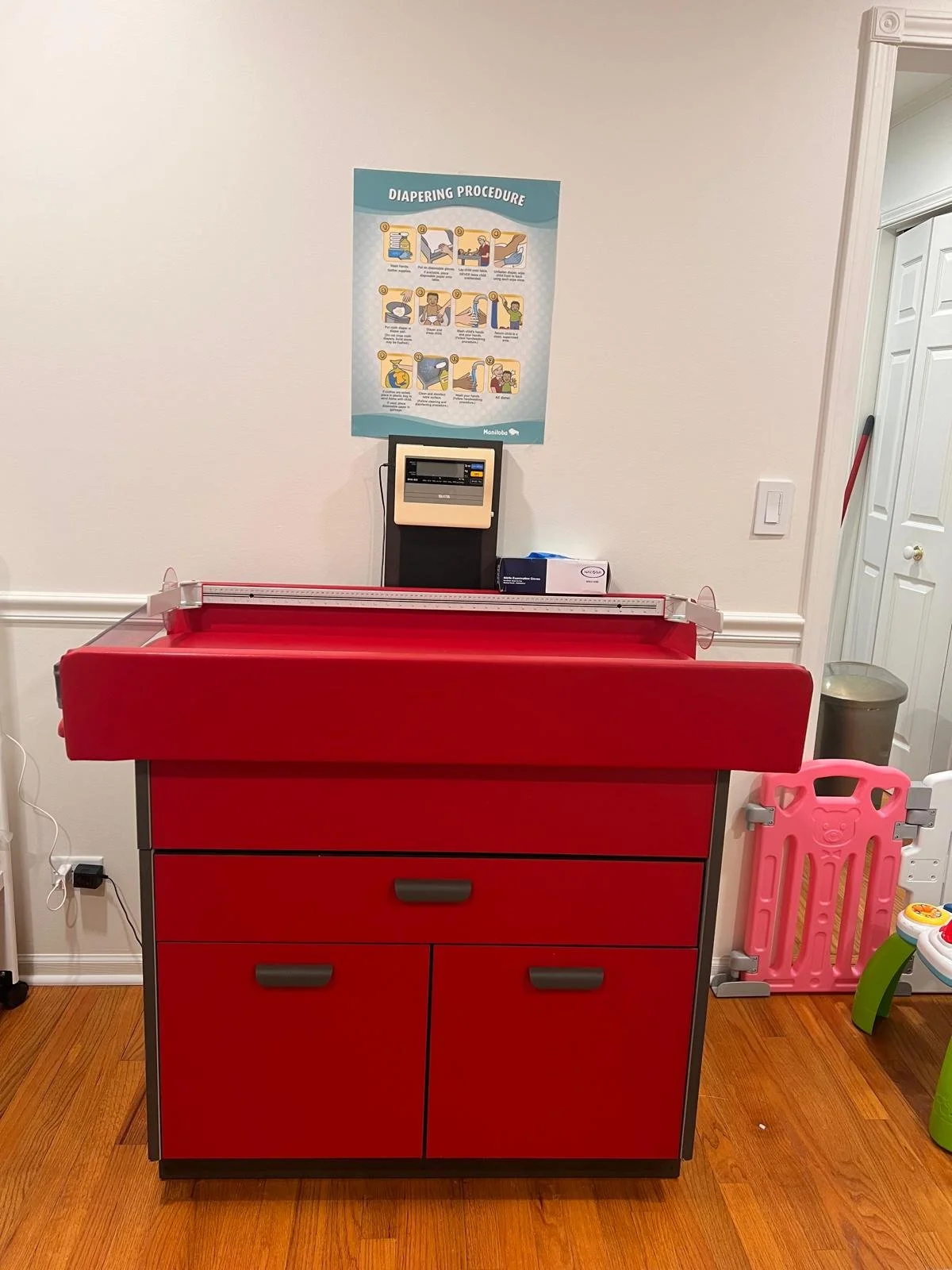 Red baby changing station with a diapering procedure chart above it on a wall, in a room with wooden flooring, a pink safety gate, and a plastic toy chair to the right.
