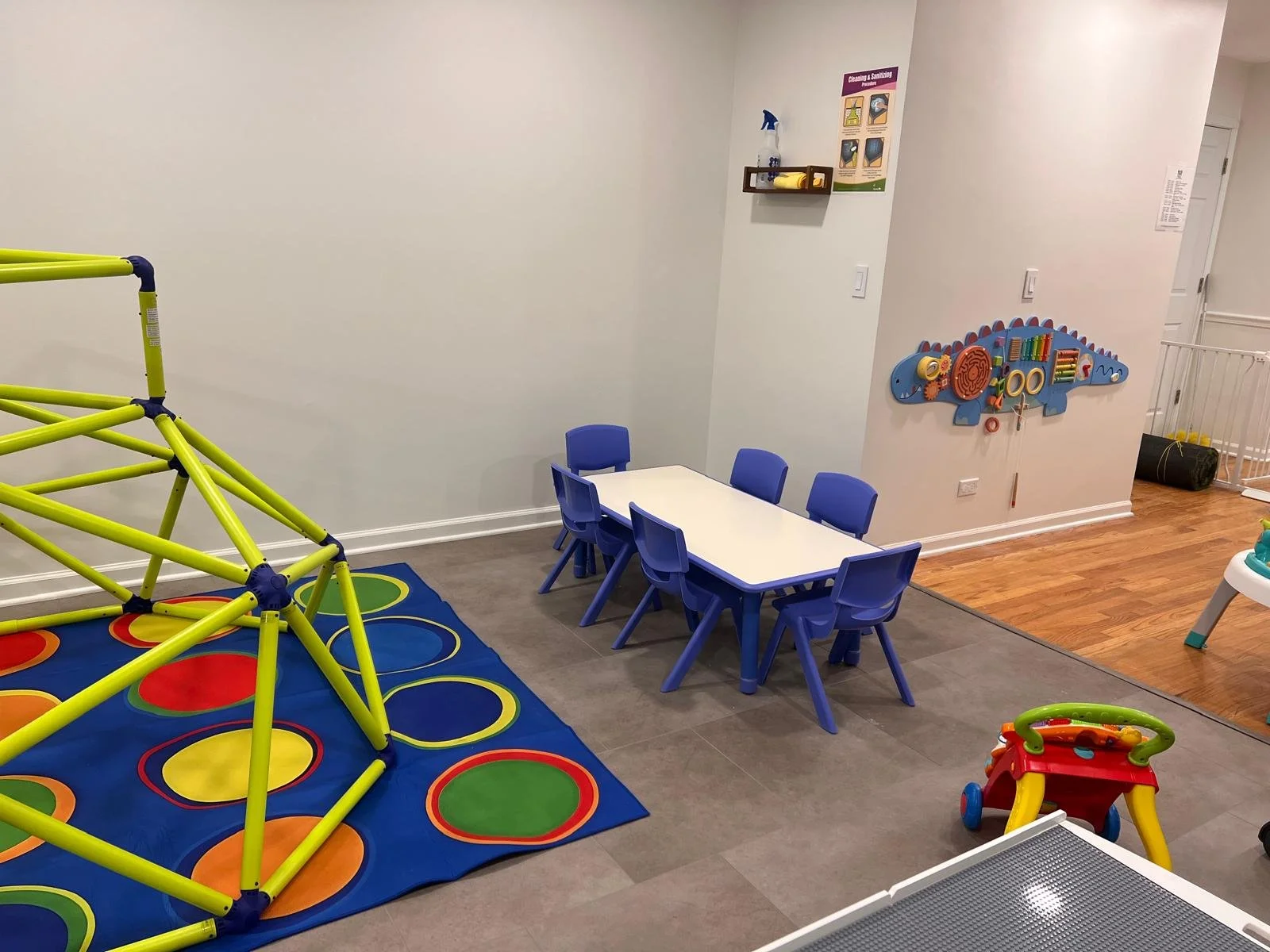 Children's play area with a colorful hopscotch mat, small blue chairs and table, a small red and yellow toy wagon, and a wall-mounted blue activity board with various shapes and toys.