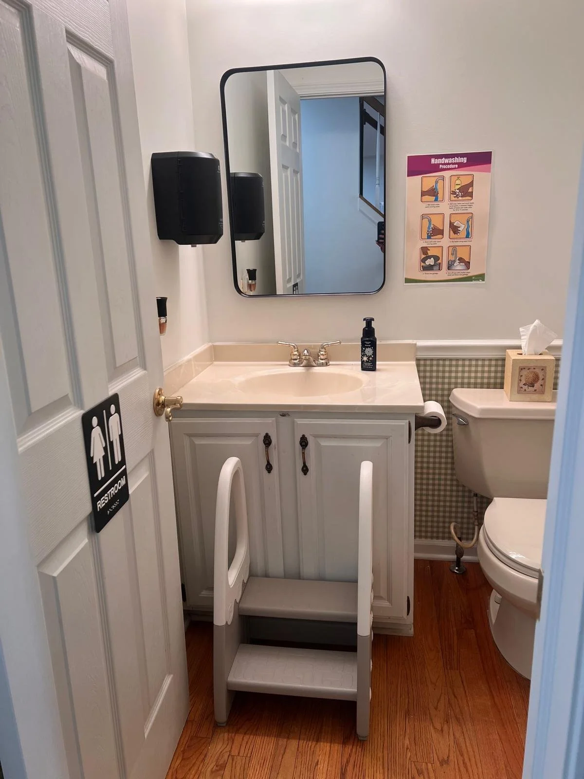 A small bathroom with a mirror, sink, and toilet. There is a step stool in front of the sink, a soap dispenser on the counter, and a box of tissues on the toilet tank. There are signs on the wall, including hand washing procedures and restroom rules.