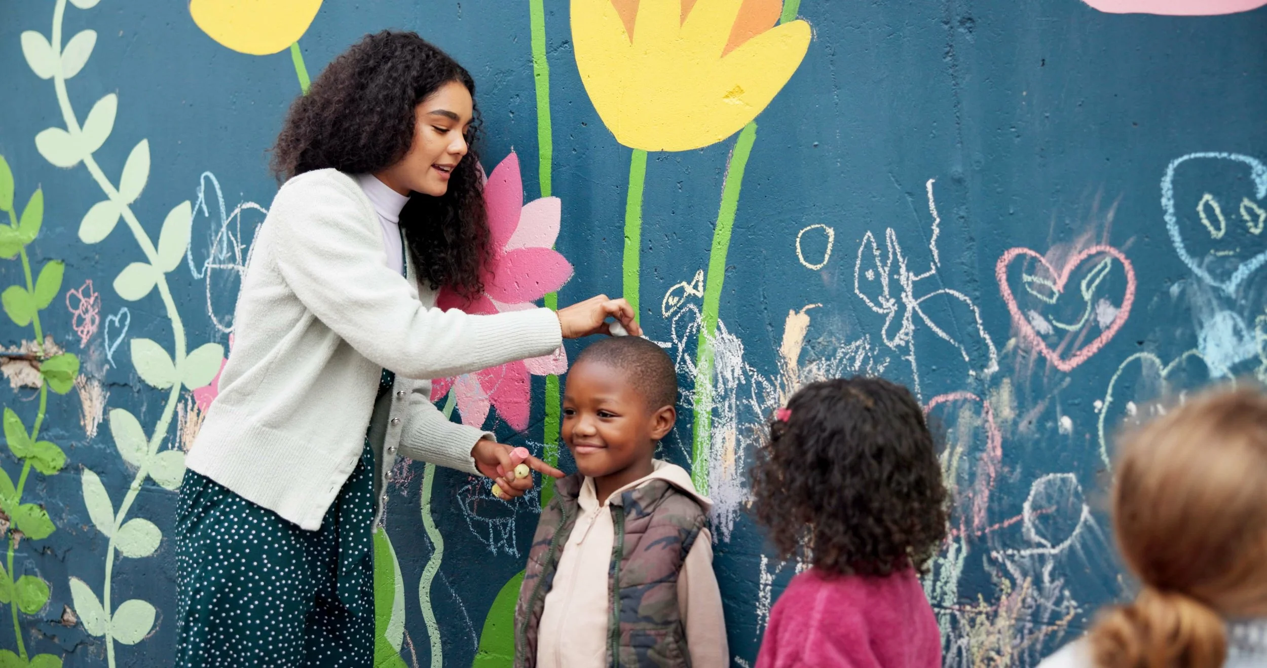 A woman drawing on a colorful chalkboard wall with children nearby. The wall features large painted flowers and various chalk drawings, including hearts and animals.